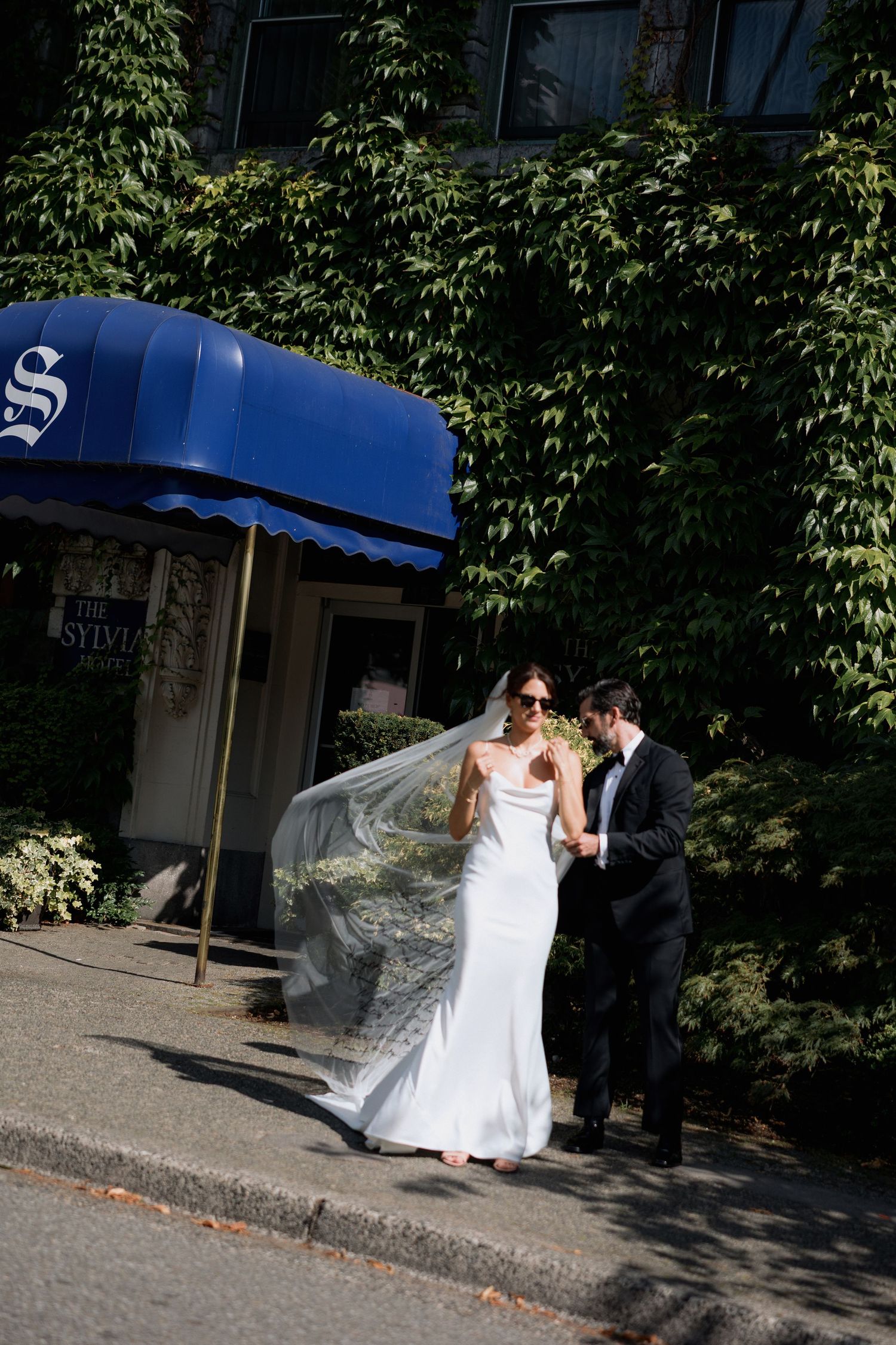 A couple in wedding attire walking together on a sidewalk near a blue storefront awning in front of the Sylvia Hotel in Vancouver