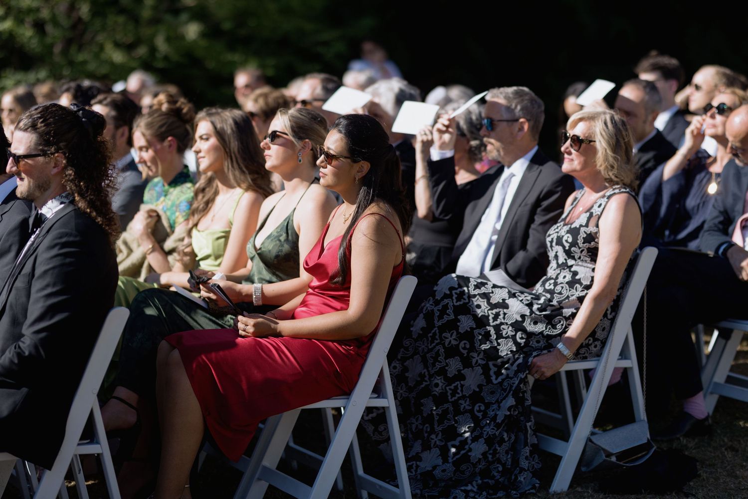 Wedding guests in colorful dresses seated in white chairs during outdoor ceremony.