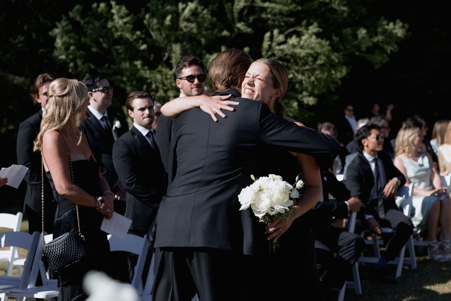 Wedding attendees stand in formal attire during an outdoor ceremony surrounded by trees.