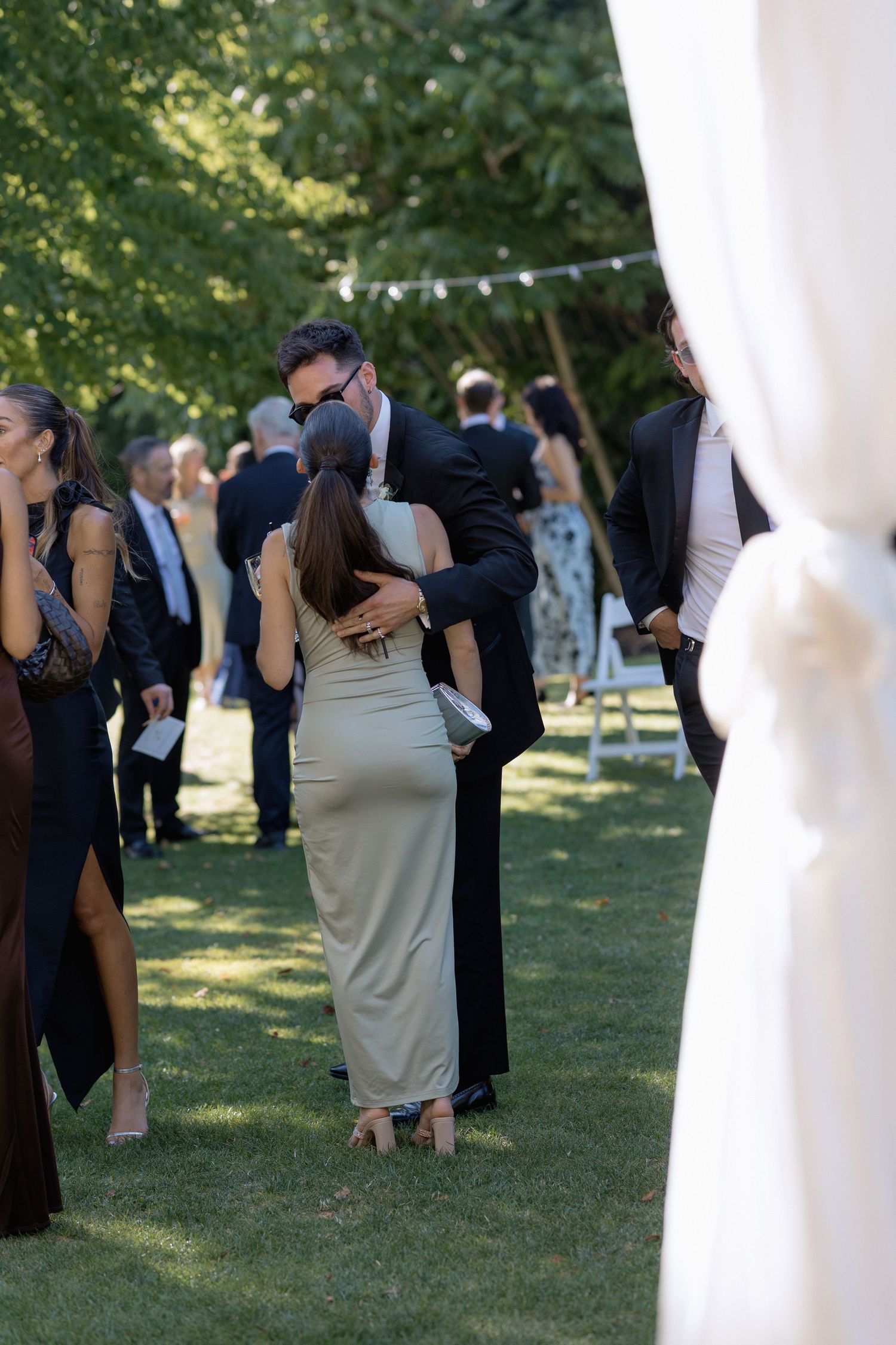 Wedding reception guests dancing and mingling on a sunlit lawn with string lights overhead.