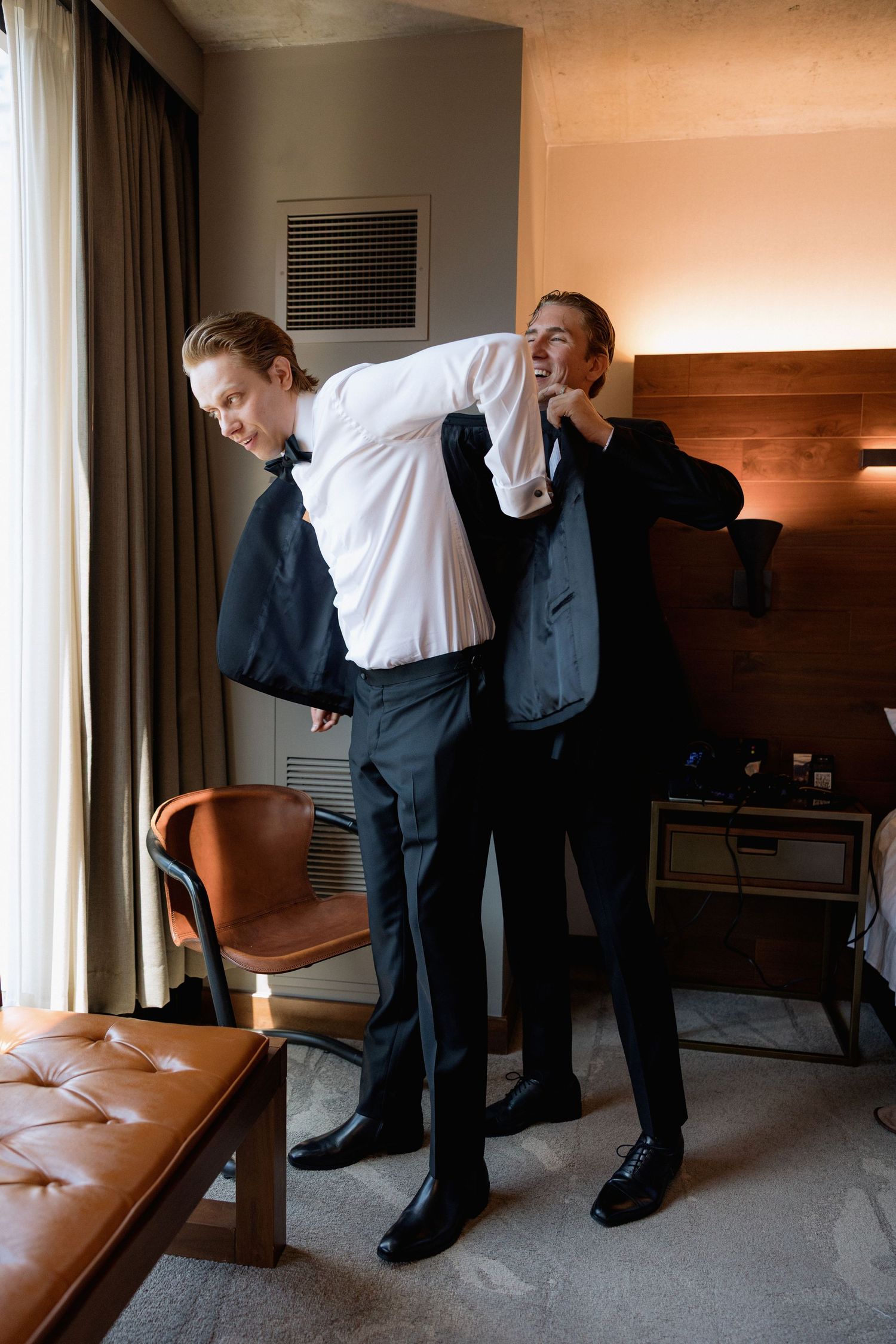 Men in formal attire playfully posing in a modern hotel room.