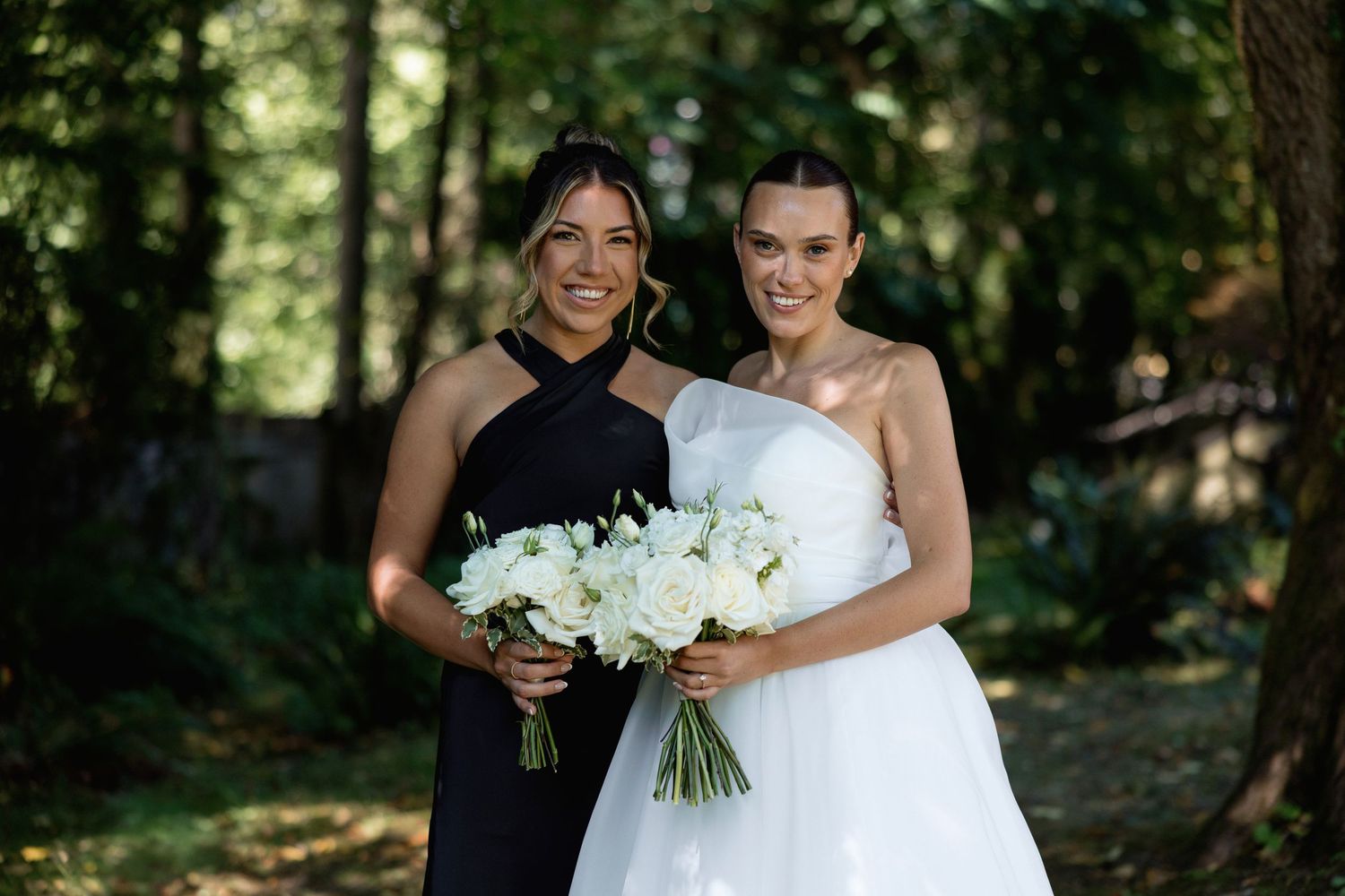 Joyful wedding portrait with attendant in black and bride in white holding bouquets outdoors.