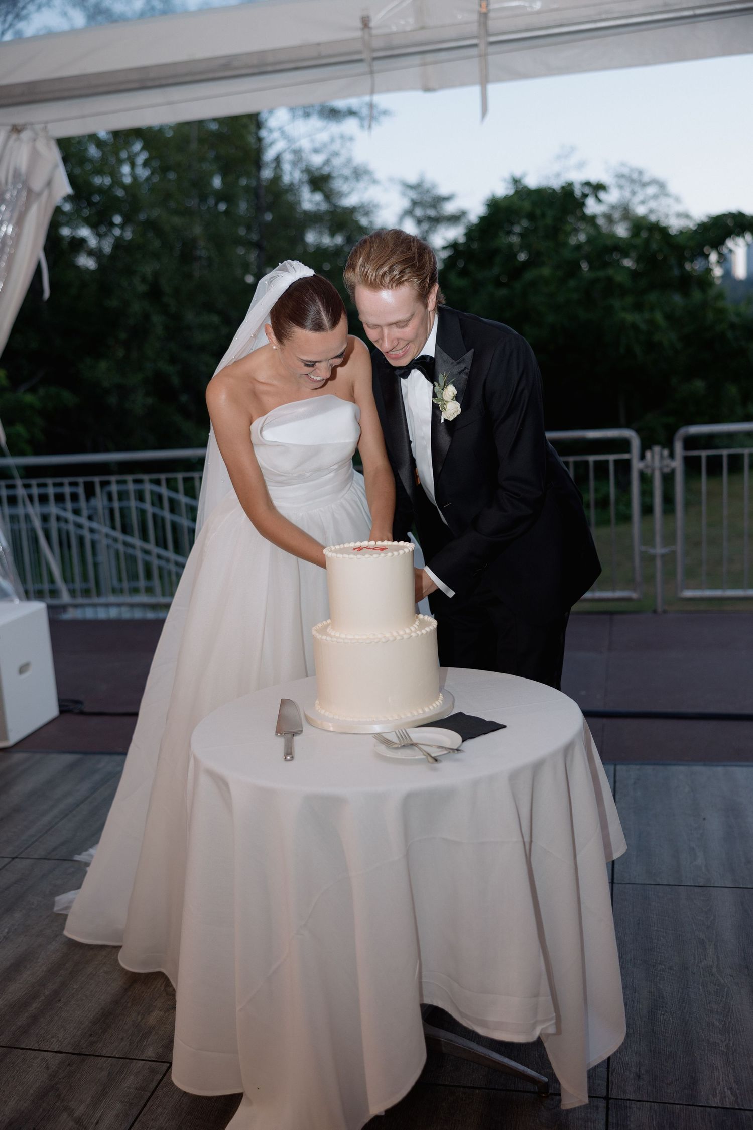 A couple in formal wedding attire examining their wedding cake on an outdoor terrace.