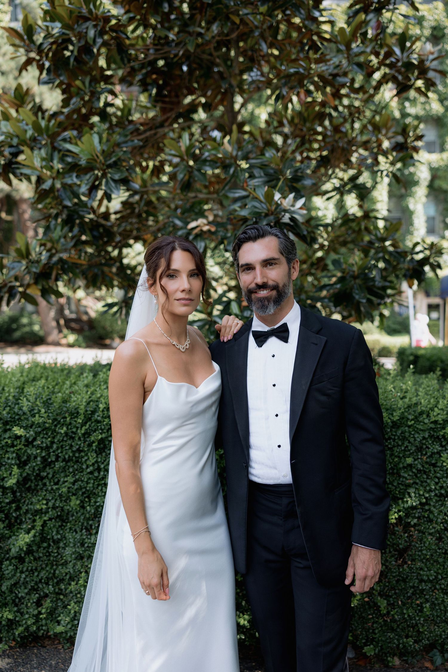 Elegant wedding couple poses in front of magnolia trees wearing classic white gown and black tuxedo.