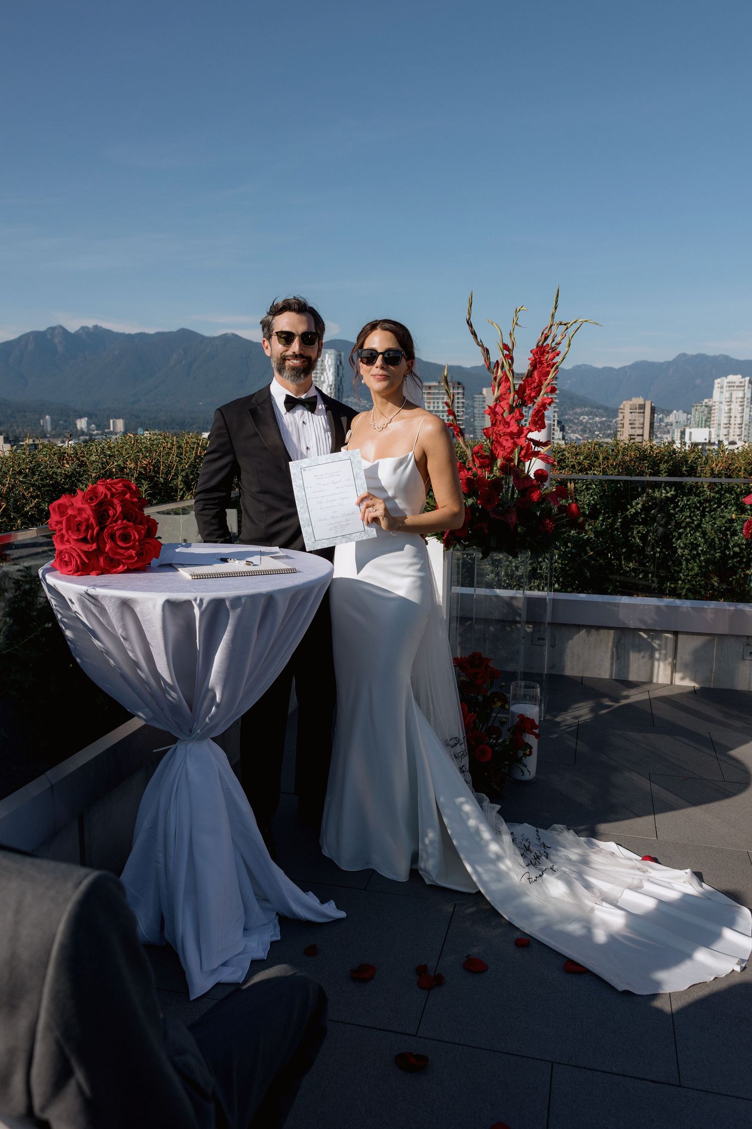 Elegant rooftop wedding setup with white linens and red floral accents against urban backdrop.