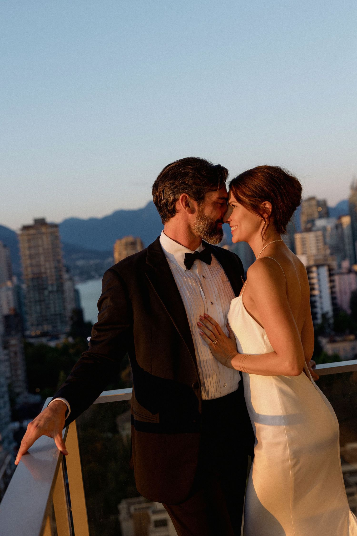 Romantic couple in formal attire share an intimate moment on a balcony overlooking a cityscape at sunset.