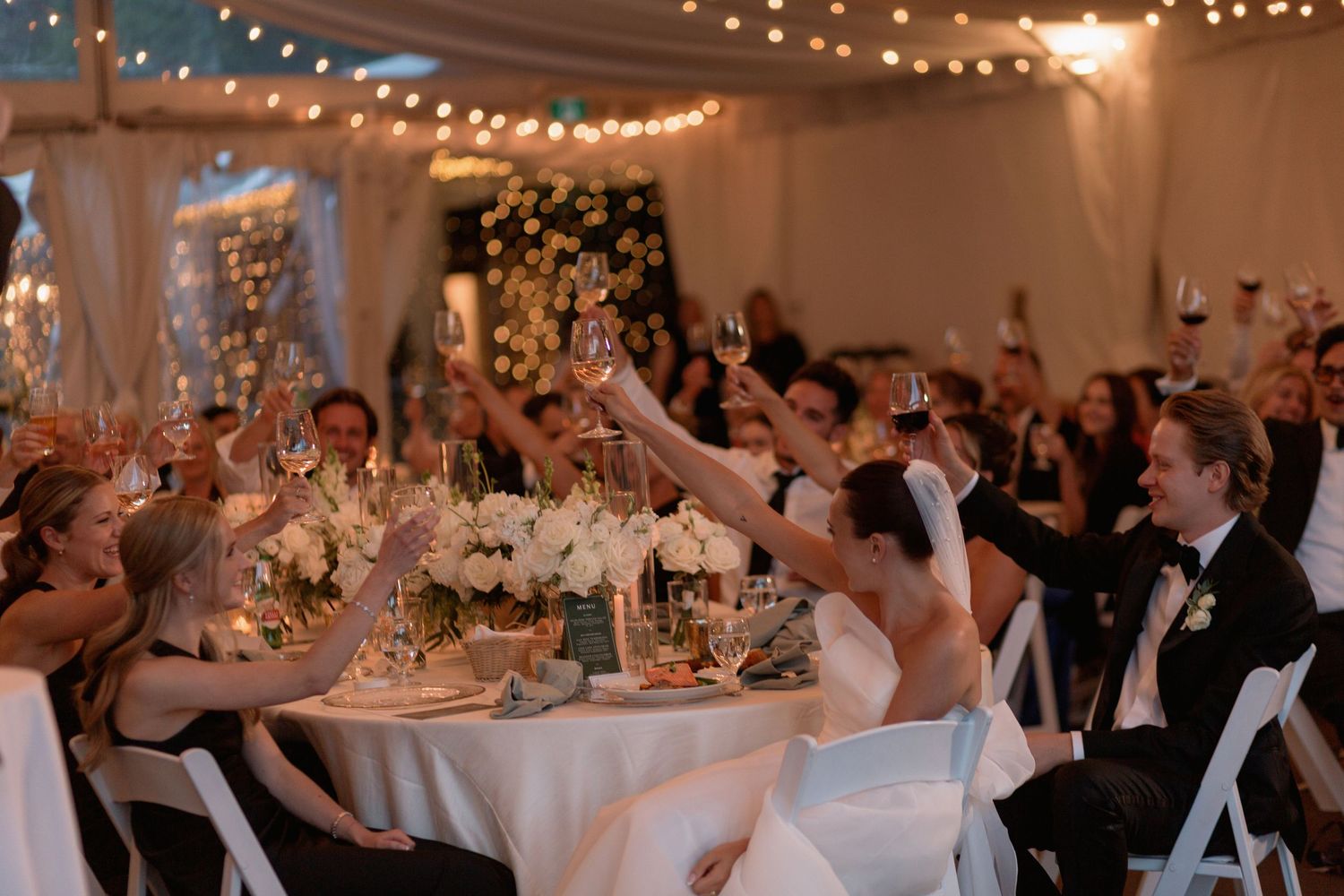 Wedding celebration under string lights with guests raising glasses in joyful toast.