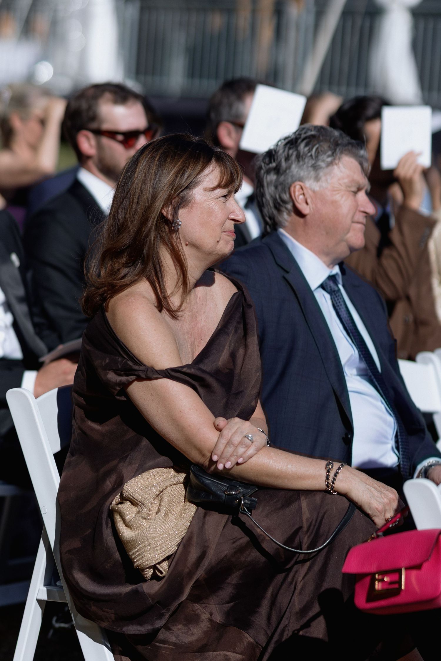 Wedding guests seated in white chairs with woman in brown dress and pink purse visible.