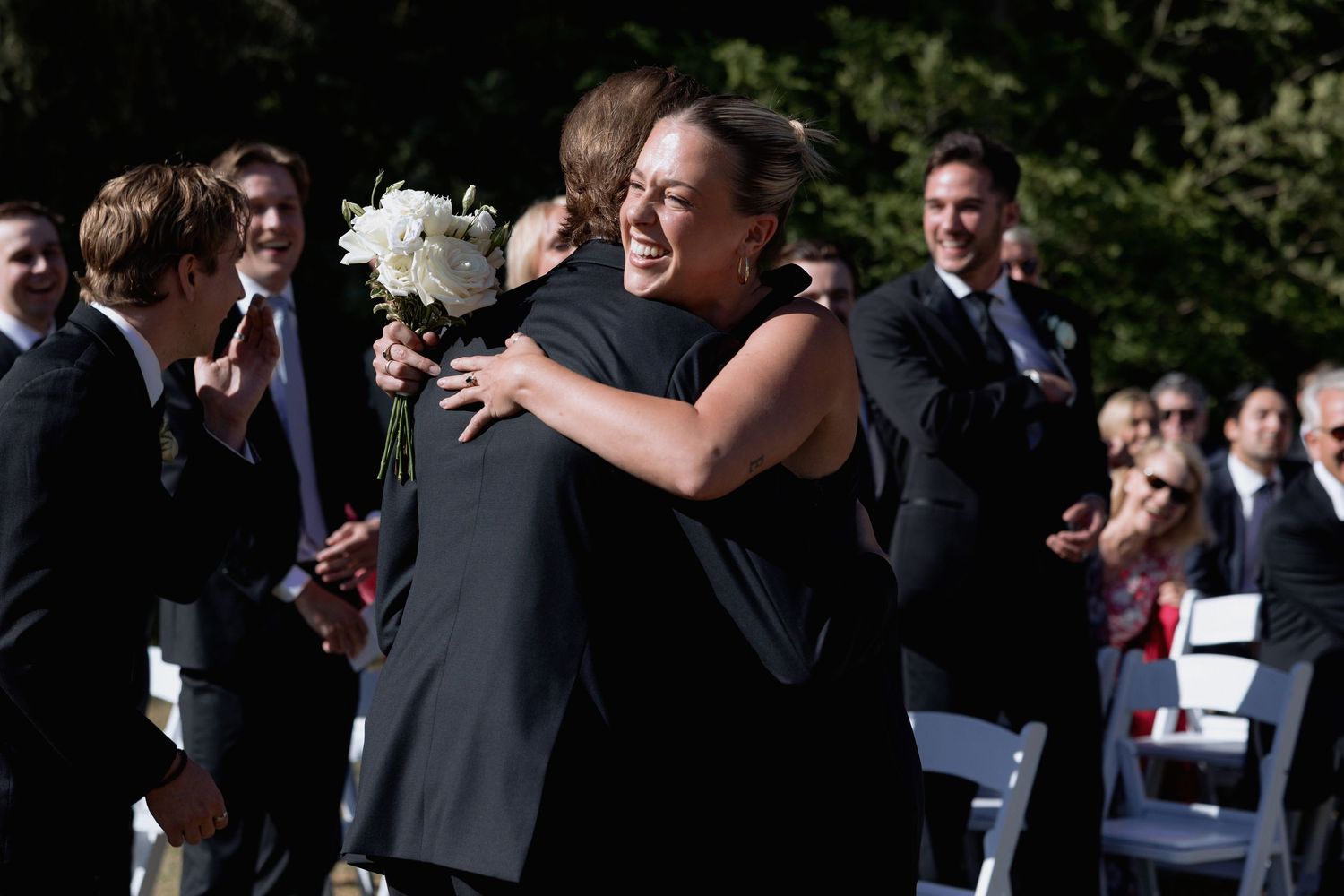 Outdoor wedding guests interact before ceremony begins in a wooded setting with white chairs.