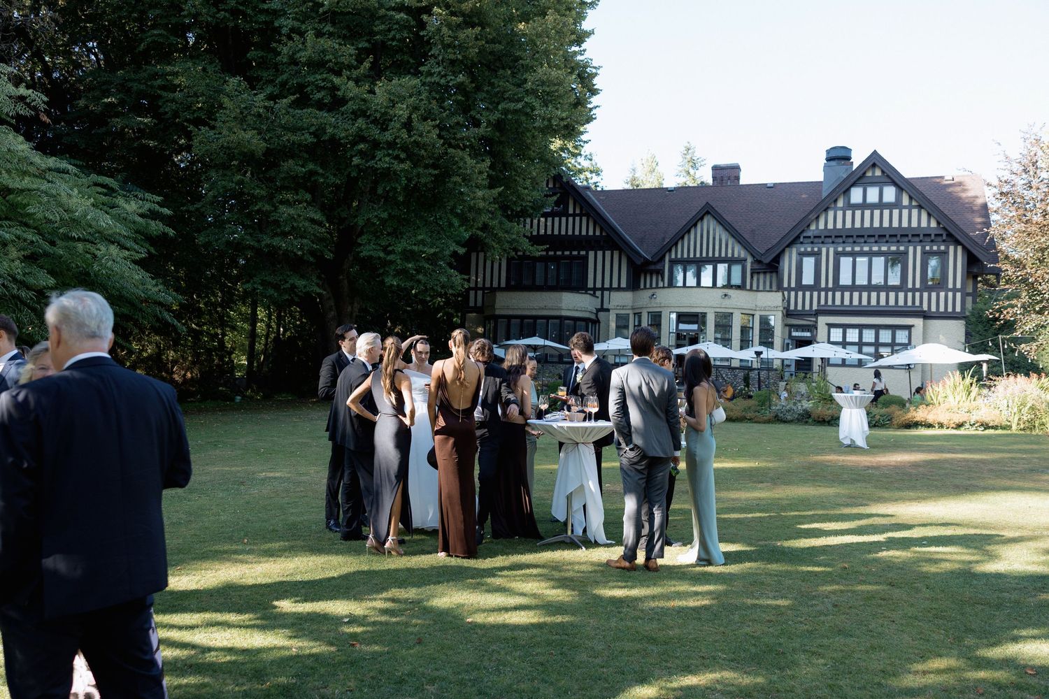 Wedding guests gathered on a lawn in front of a Tudor-style mansion during an outdoor reception in Hurt House in Burnaby