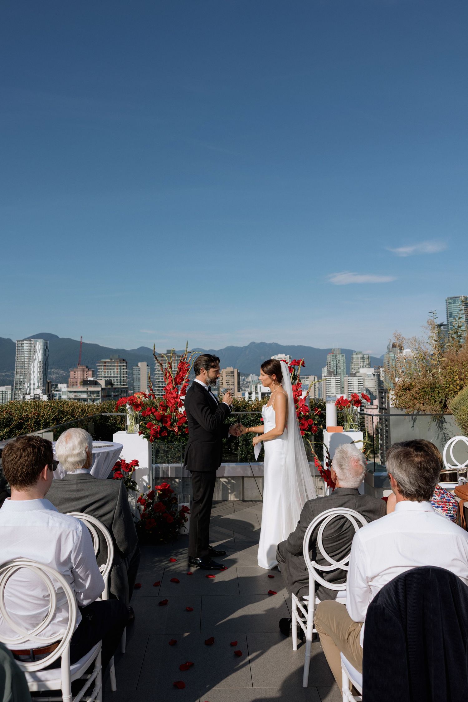 Rooftop wedding ceremony in Vancouver with red floral arrangements and white chairs against city skyline backdrop.