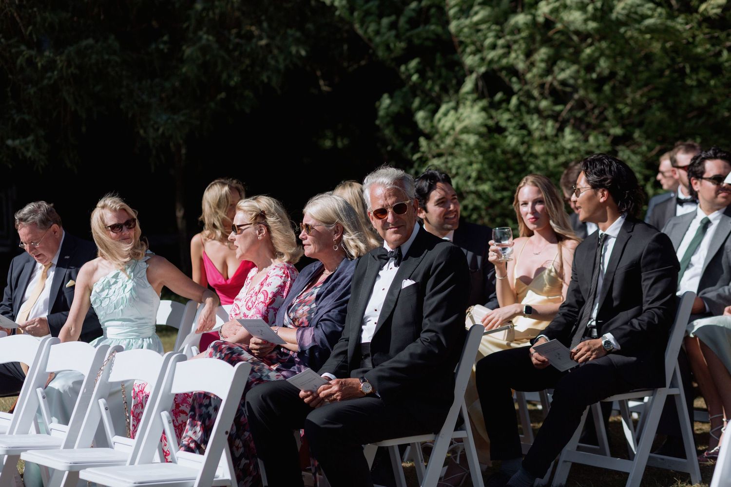 Guests in formal attire seated in white folding chairs at an outdoor wedding.