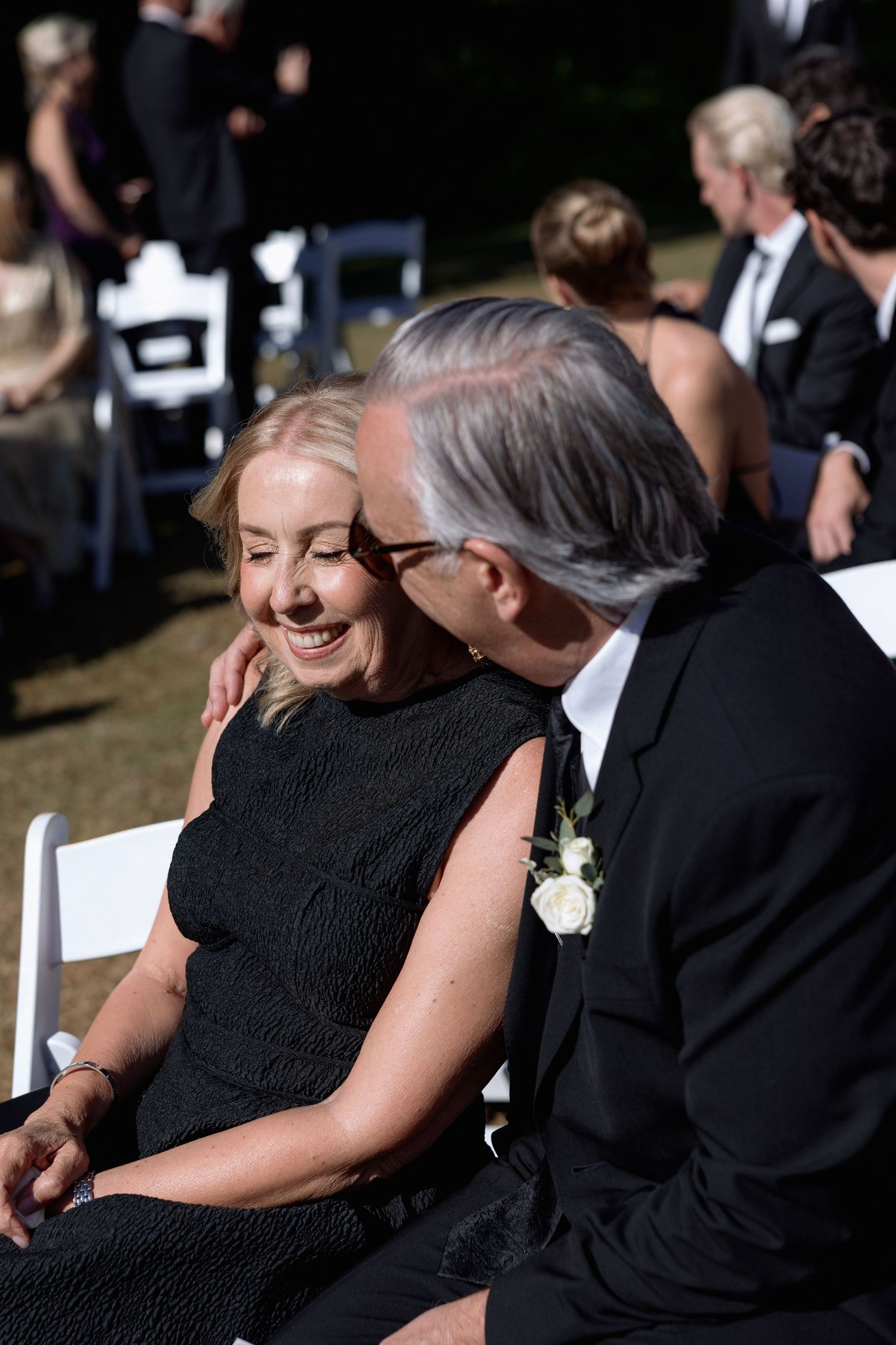 Guests in formal black attire share an intimate moment during an outdoor wedding ceremony.