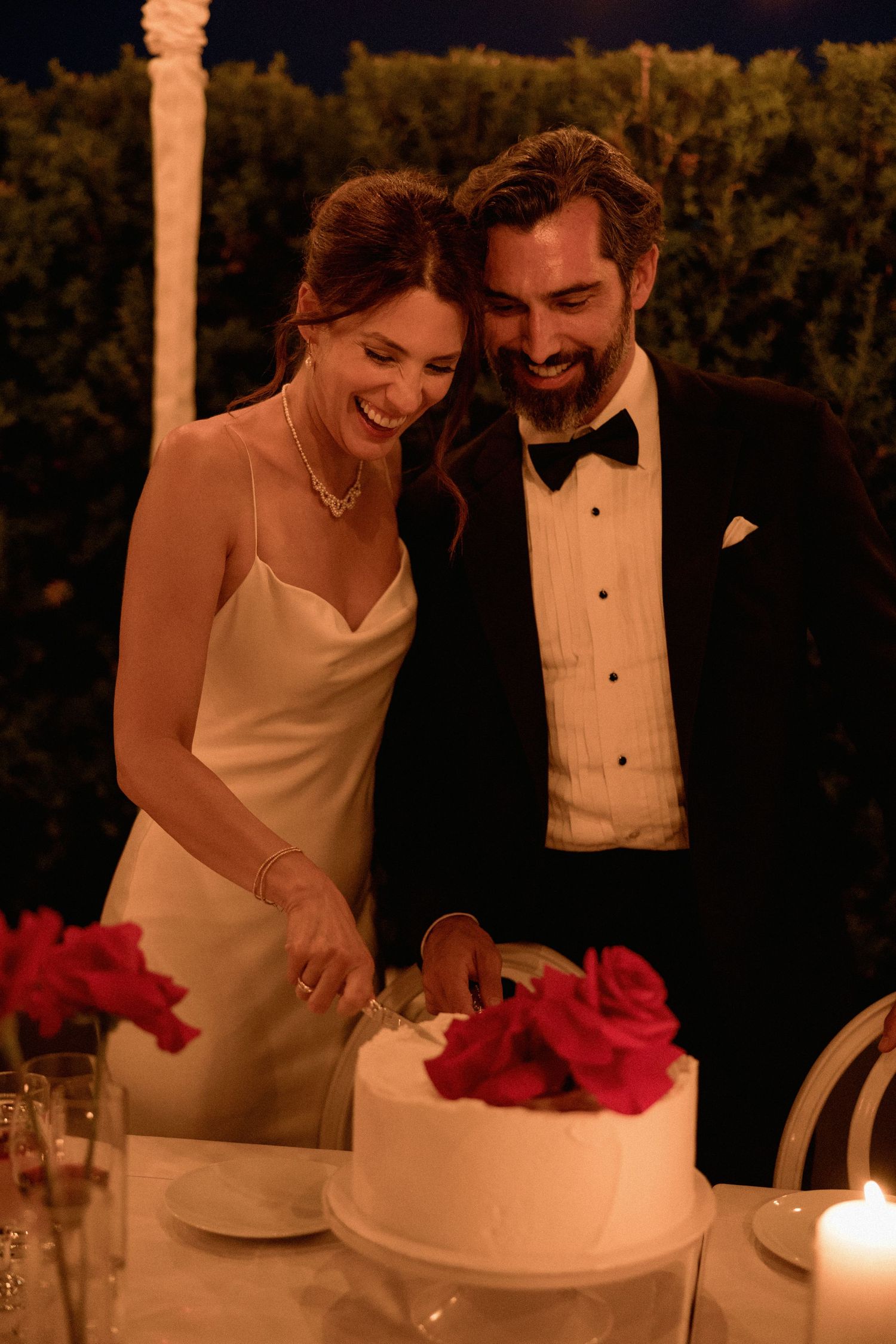 Newlyweds sharing a joyful moment while cutting their white wedding cake decorated with red roses in Vancouver