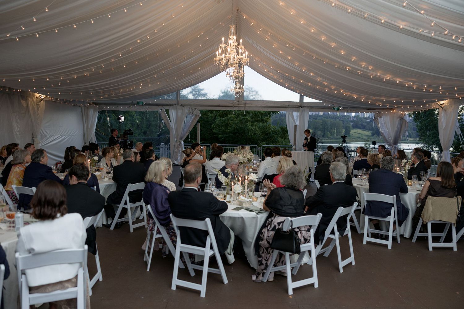 Wedding reception in white tent with crystal chandeliers and folding chairs around circular tables.