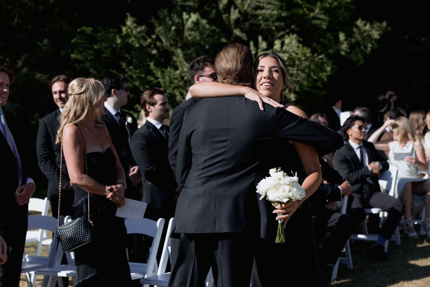 Wedding guests in formal wear socialize at an outdoor ceremony site with white chairs.