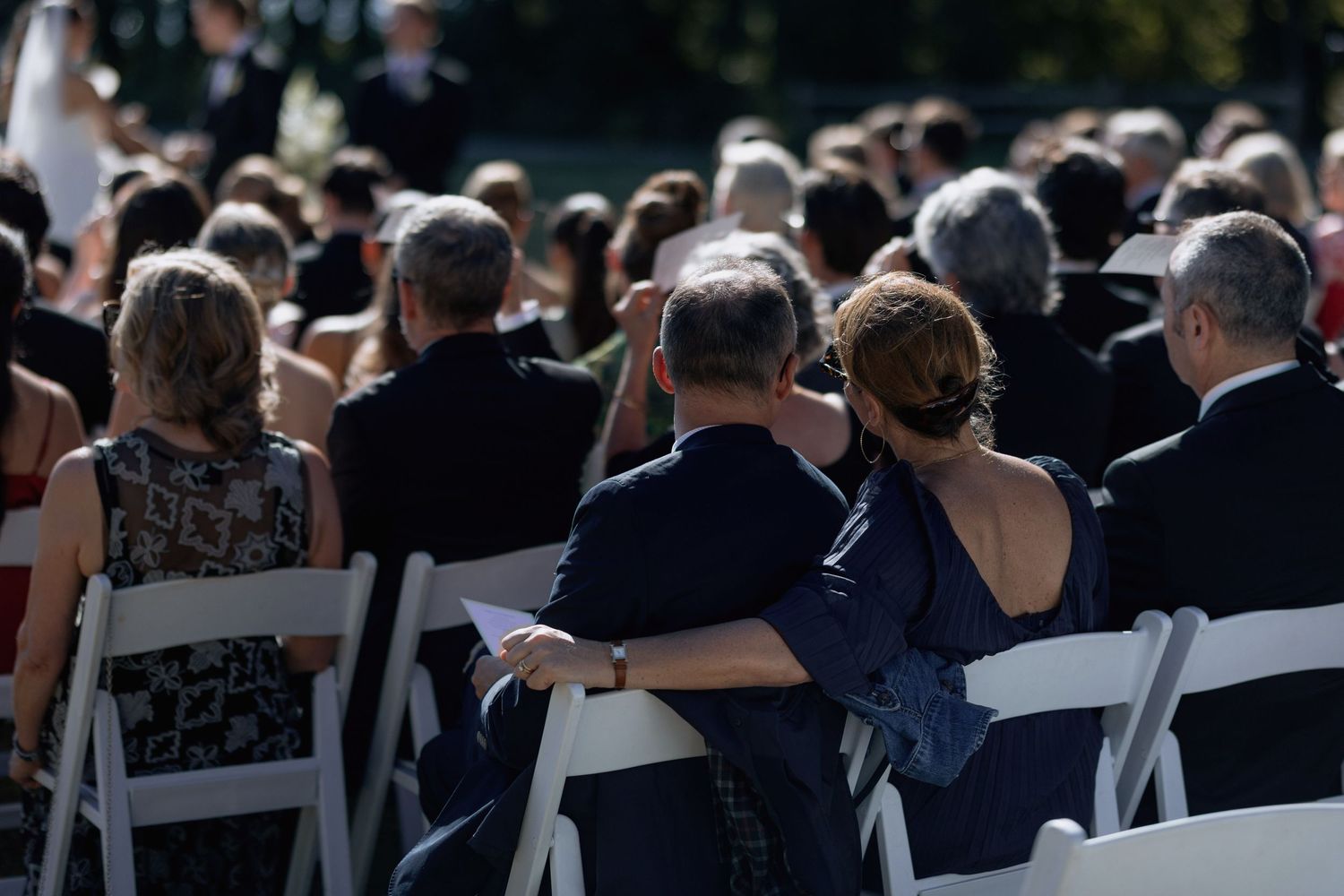 Wedding guests seated in white chairs at outdoor ceremony viewed from behind.
