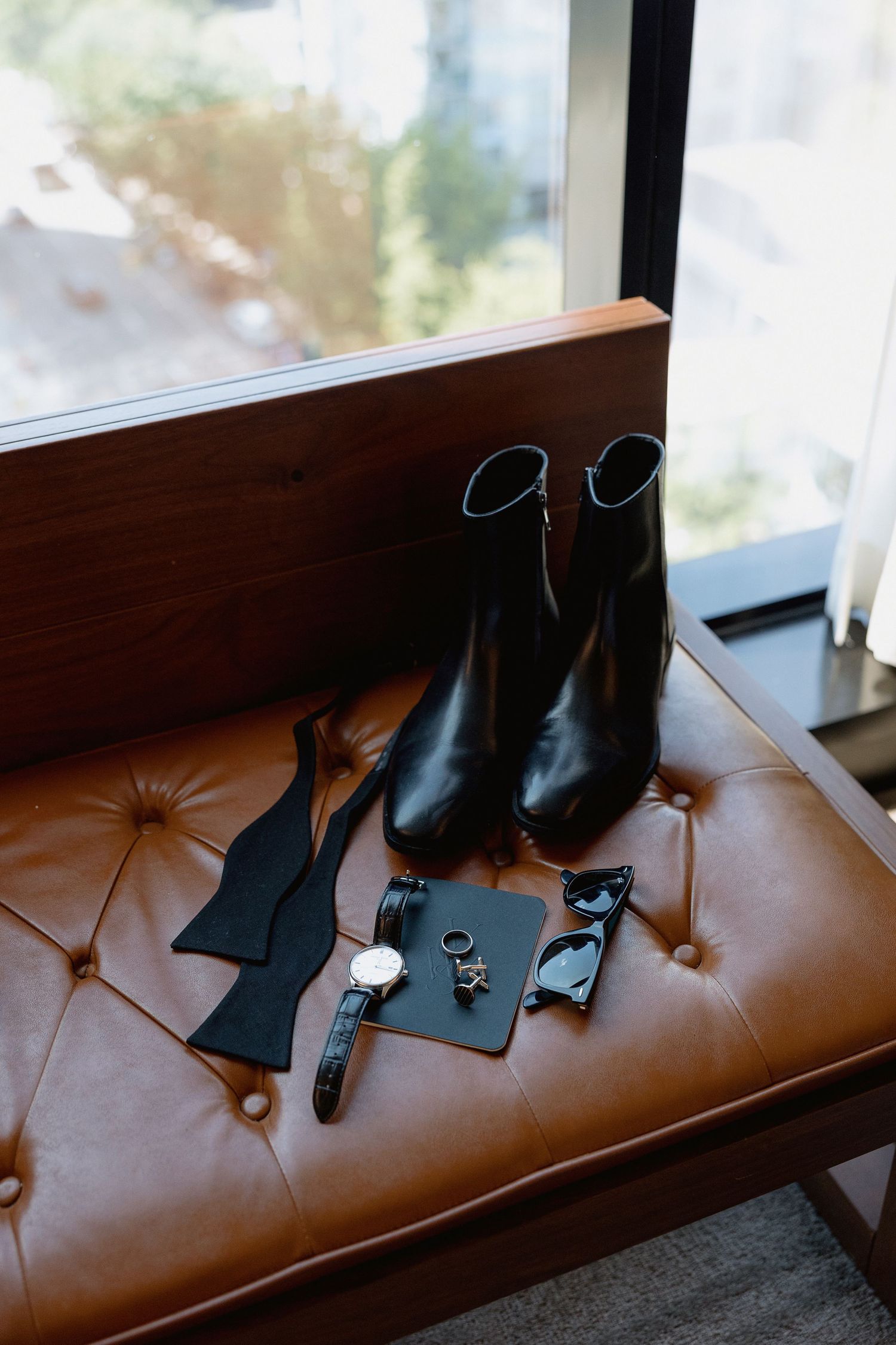 Black formal dress shoes arranged on a brown leather bench by a window.