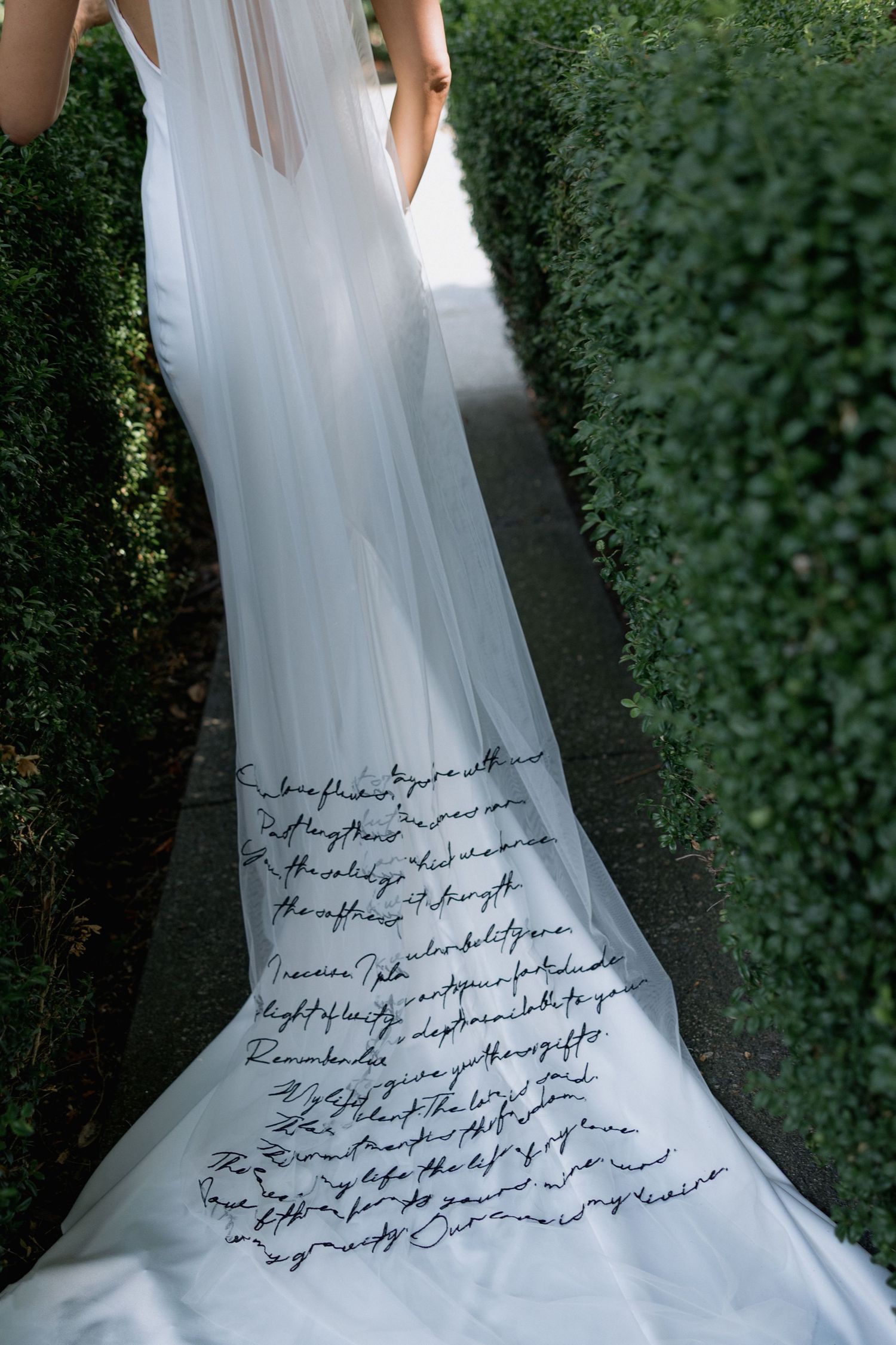 A white wedding dress with handwritten text embroidered on the train flows between green garden hedges.