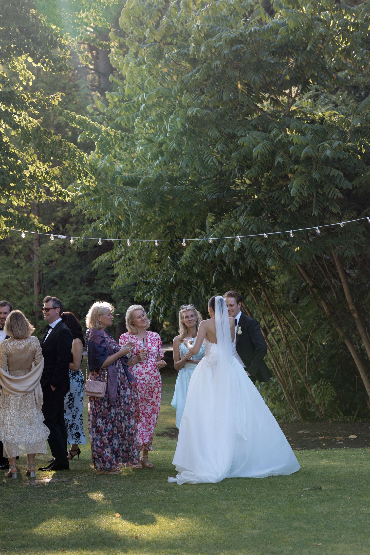 Wedding guests gather on a lawn with string lights and trees in the background.