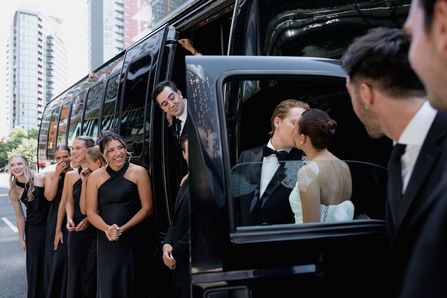 Wedding party poses playfully while entering a black limousine on a city street.
