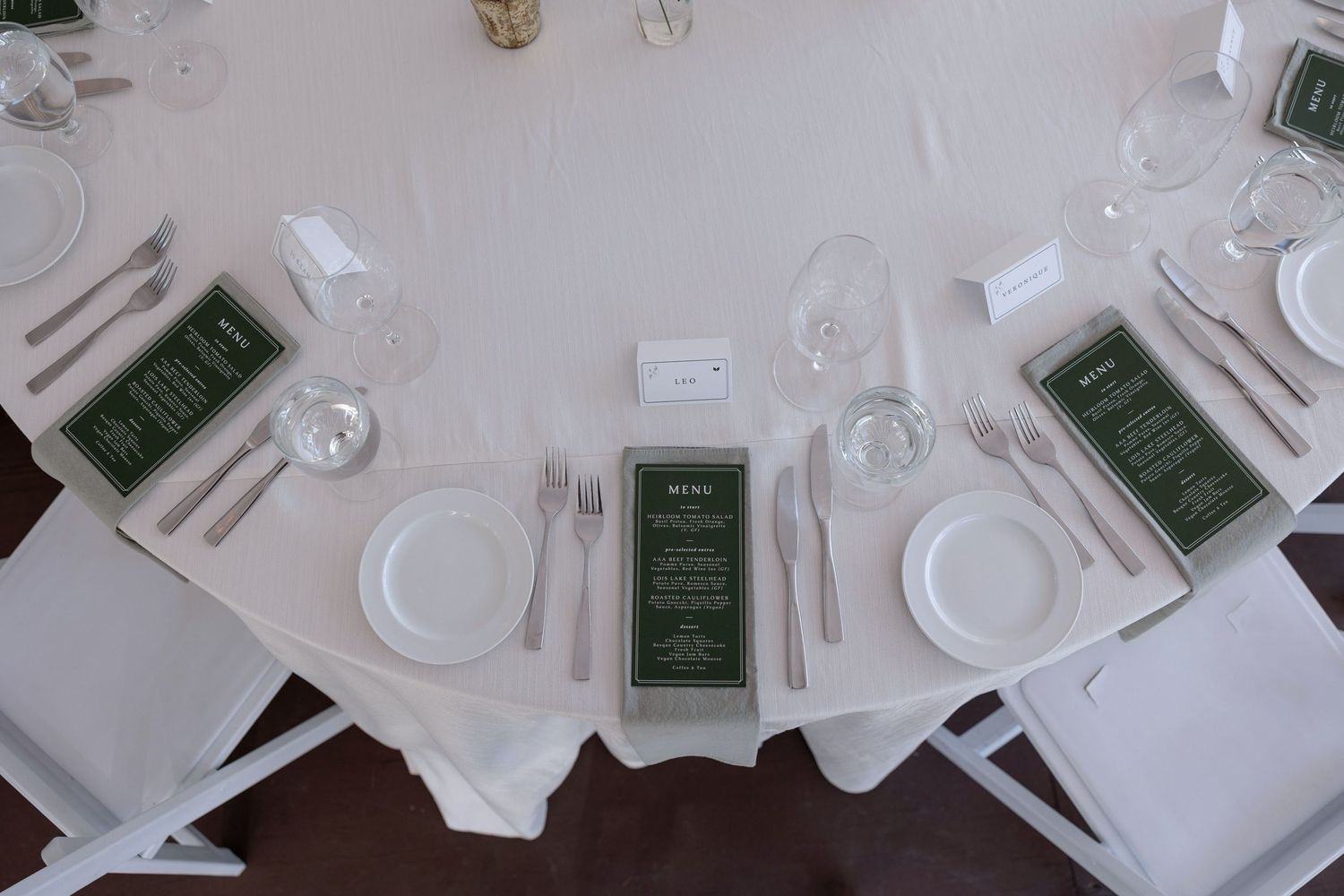 Close-up of formal place settings with white plates, glassware and green menu cards on round tables.