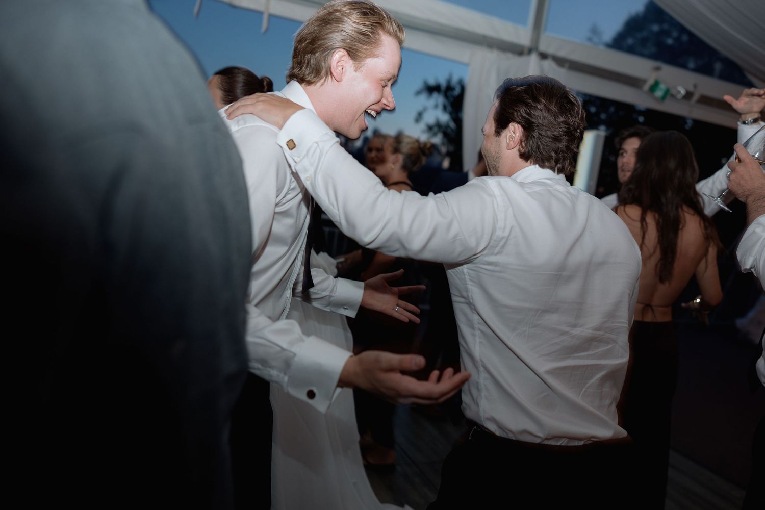 Wedding guests dance energetically under a tent with white draping and string lights.