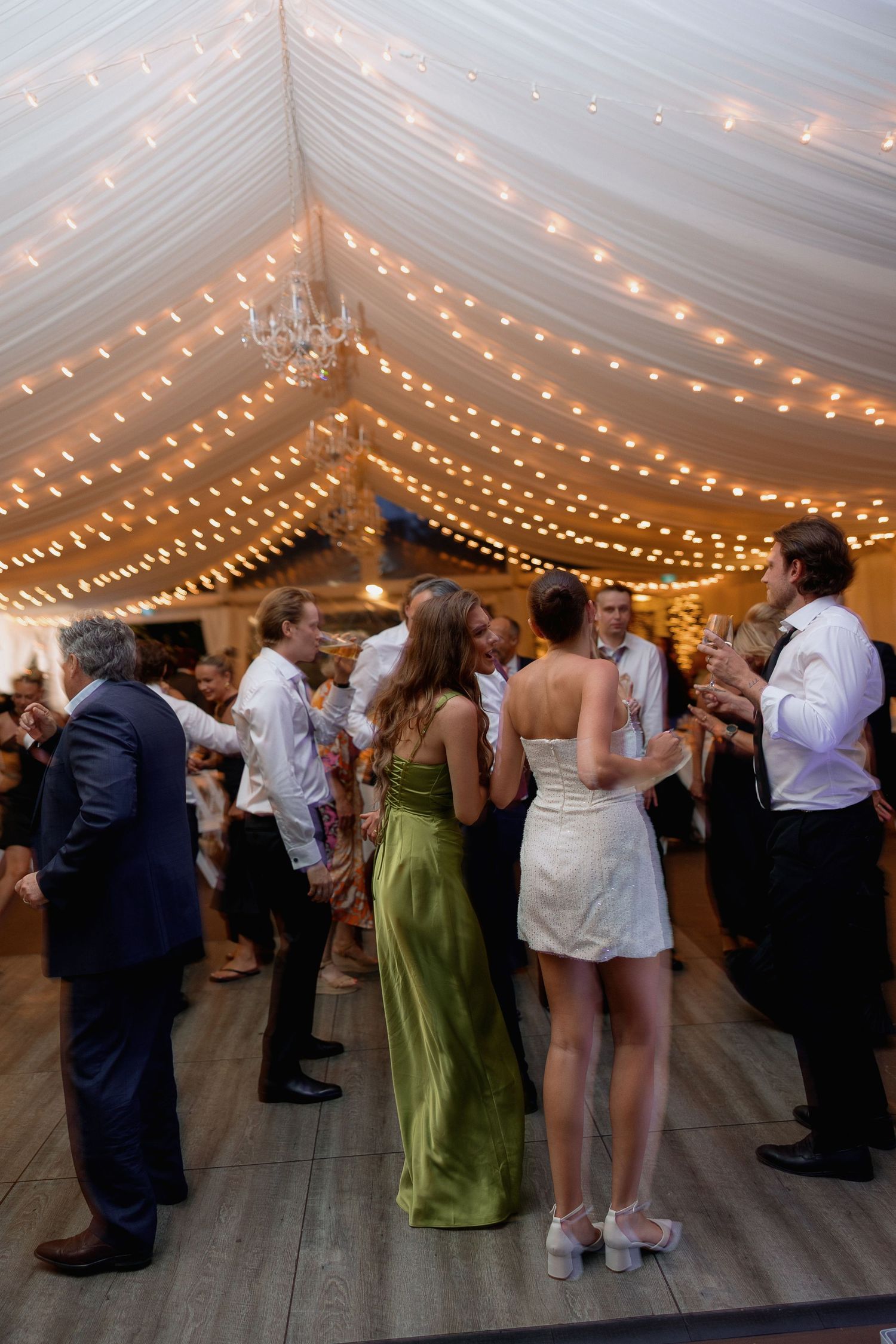 Wedding celebration in a tent with string lights as guests dance and socialize.