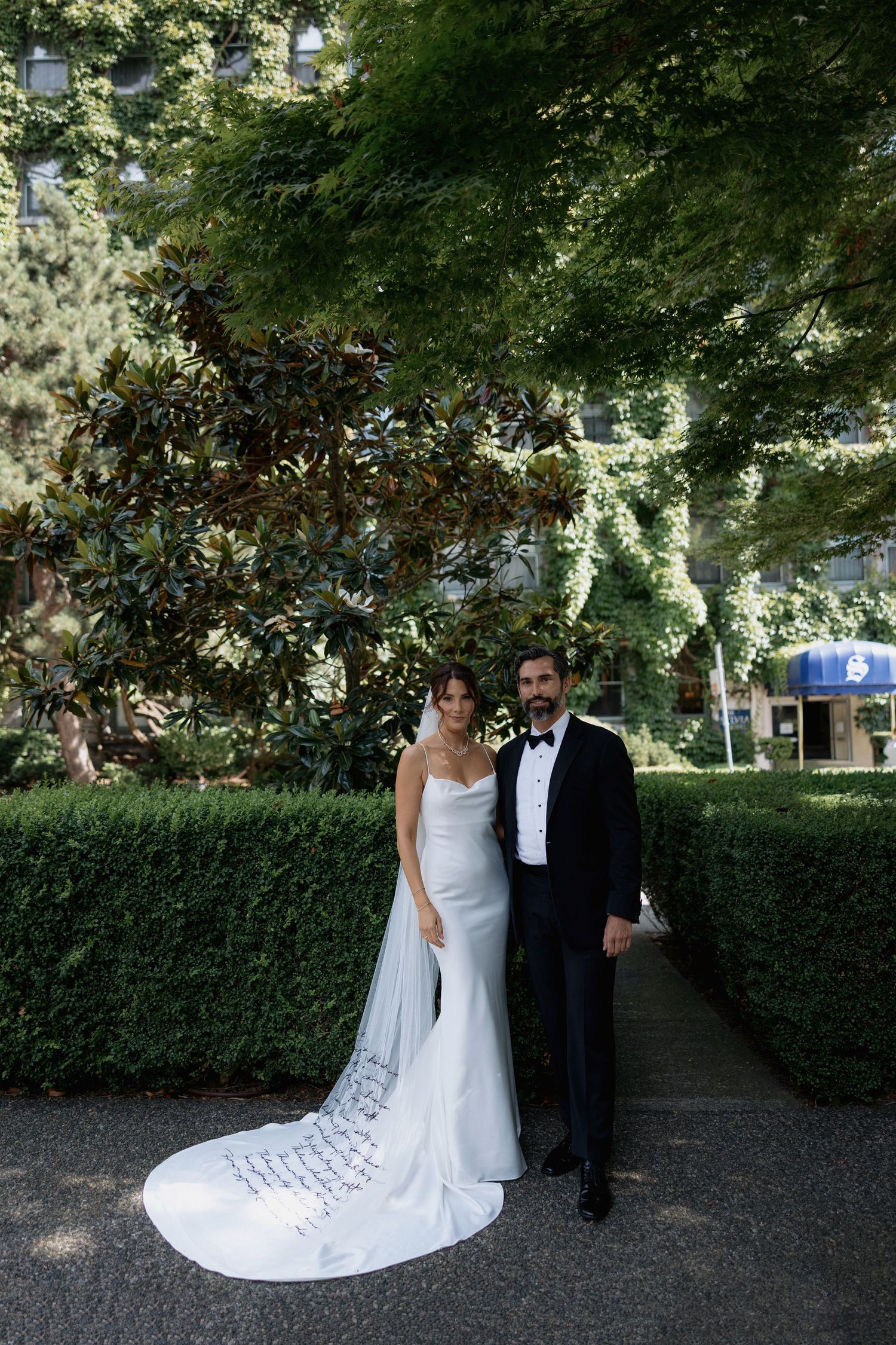 A romantic wedding couple walks together through a garden path lined with lush green hedges in front of the Sylvia Hotel in Vancouver