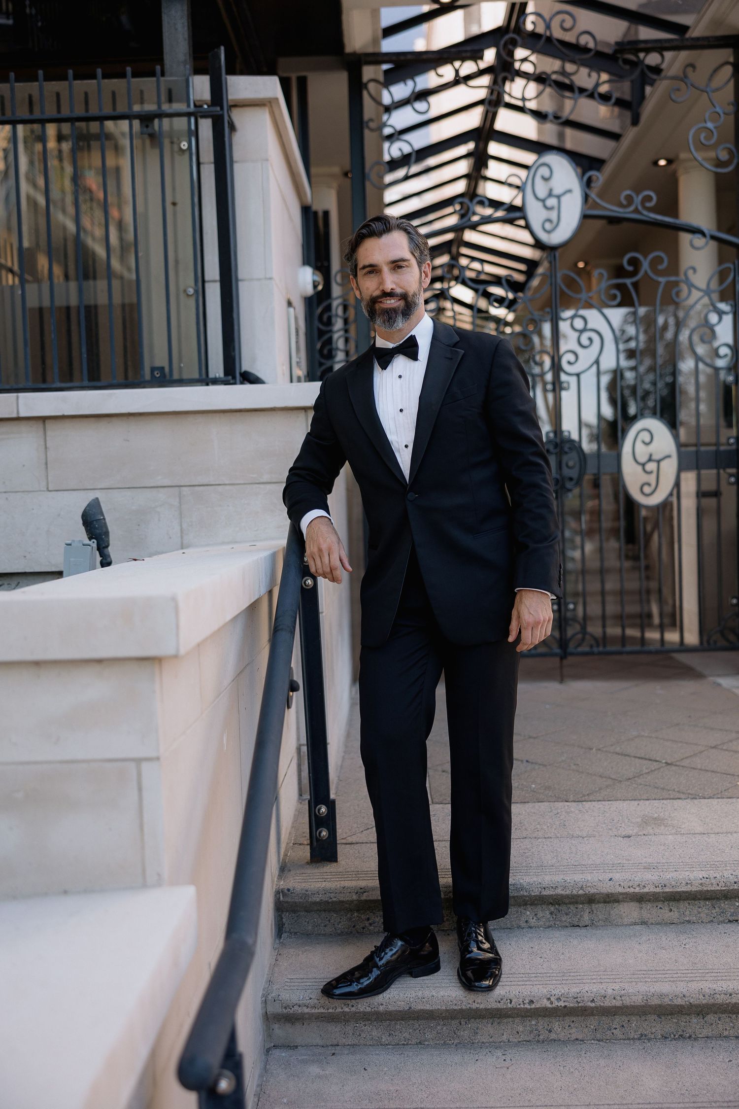 Formal portrait of person in classic black tuxedo standing on stone steps near ornate iron gates.