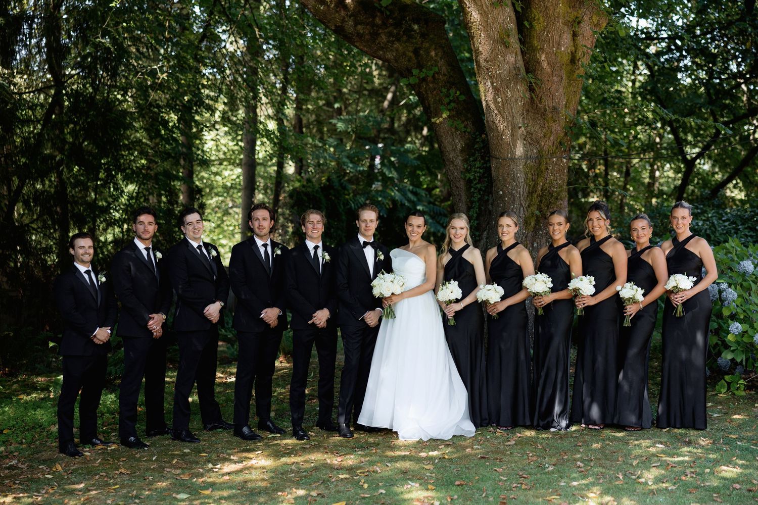 Wedding group poses in a creative formation beneath large trees in a park.