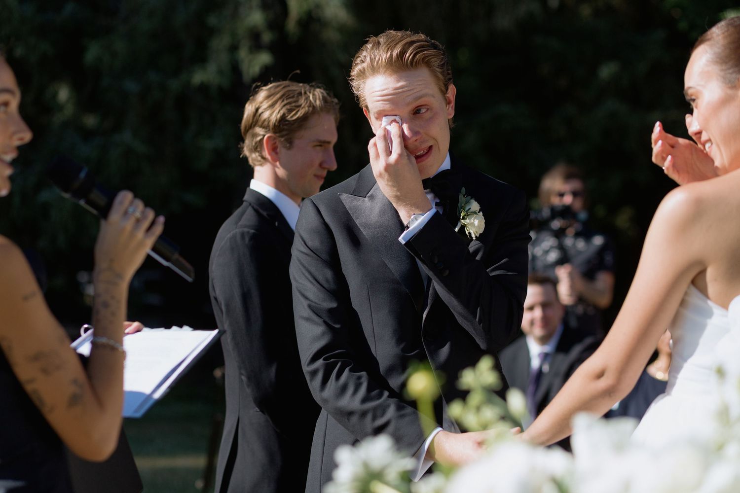 Guests seated at an outdoor wedding ceremony display emotional reactions while watching the proceedings.