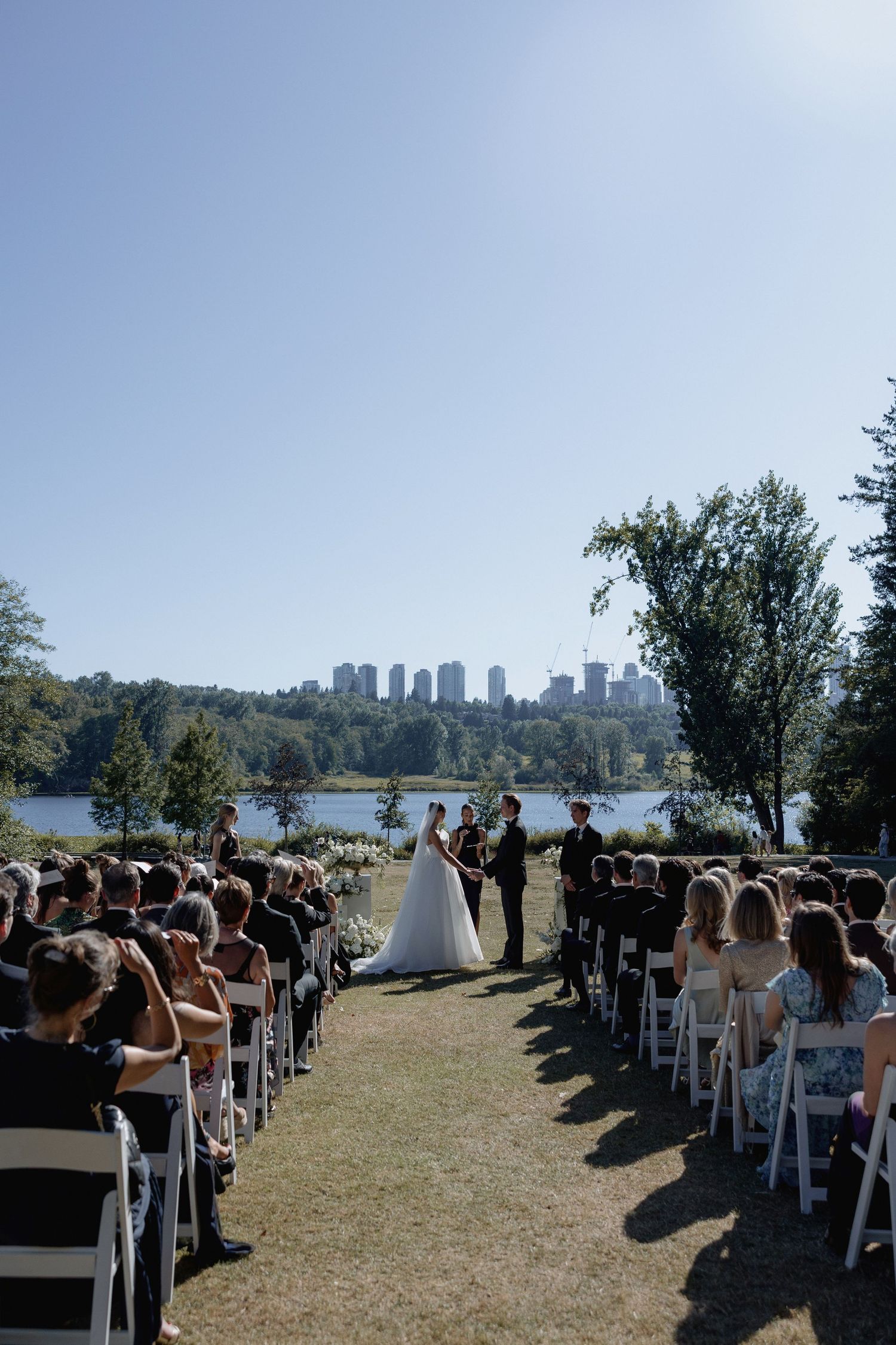 Rows of white chairs line a grassy lawn for an outdoor wedding with a city skyline in the distance and Deer Lake