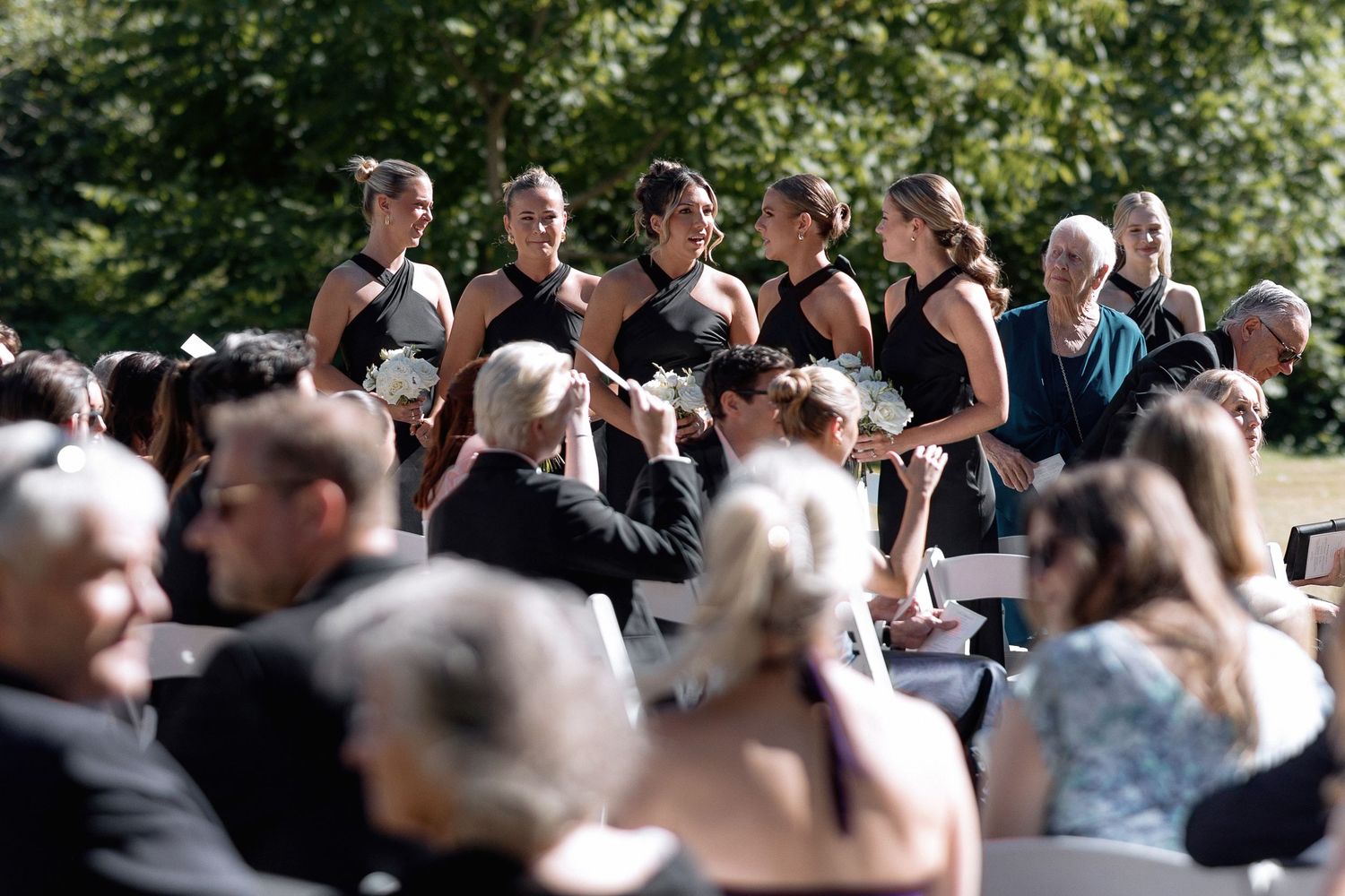 Wedding guests gather outdoors on a sunny day with trees in the background.