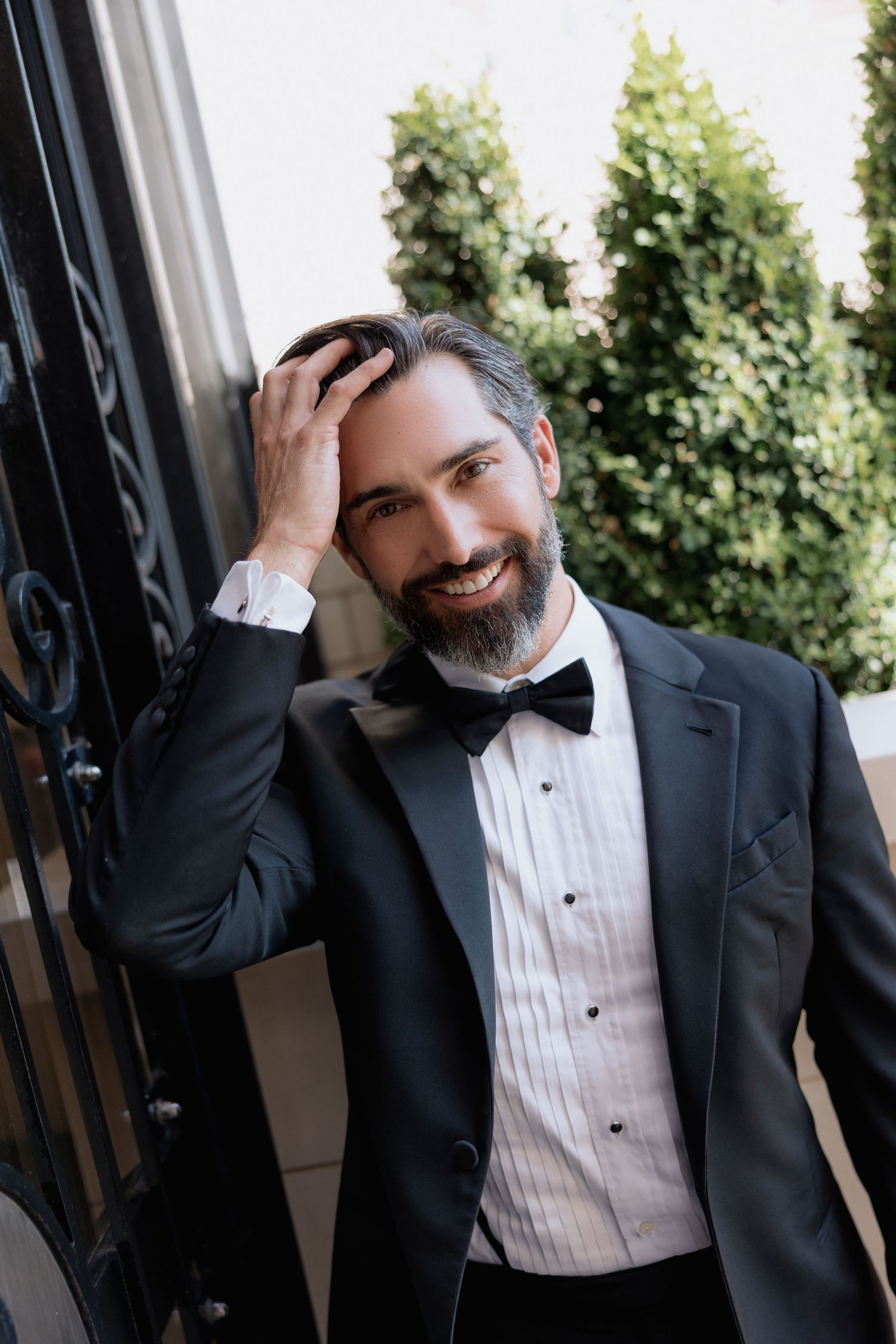 Candid moment of person in tuxedo running hand through hair while smiling near iron gate.