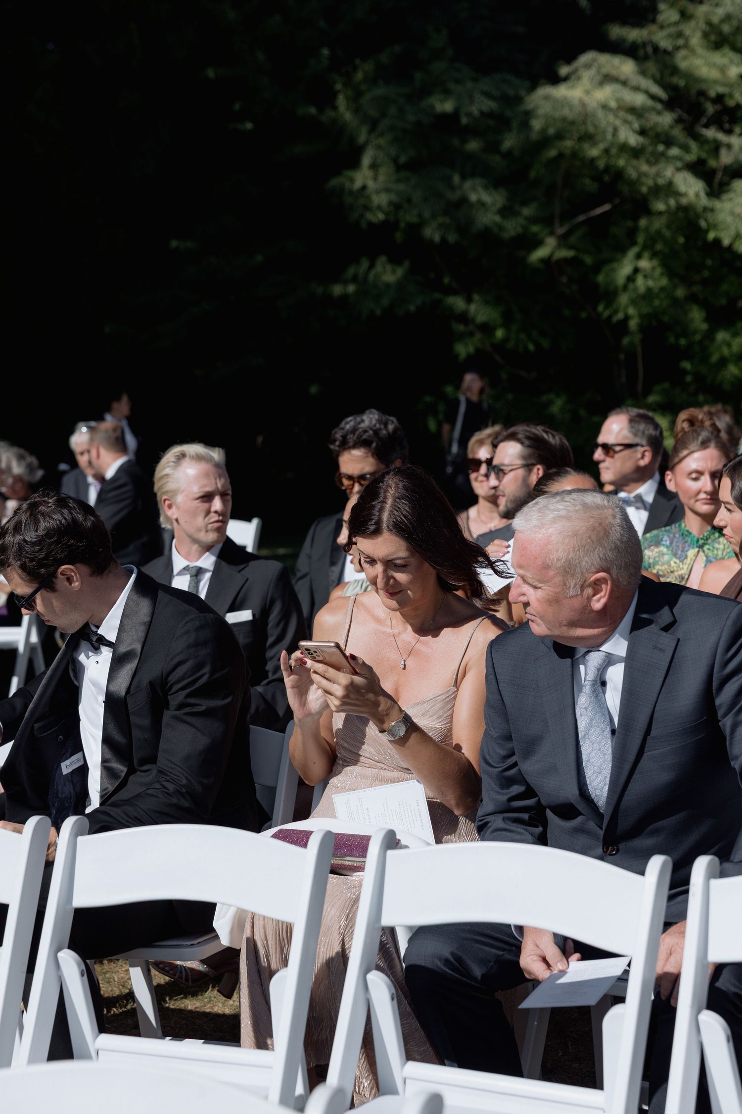Guests in formal attire seated on white chairs at an outdoor wedding venue.