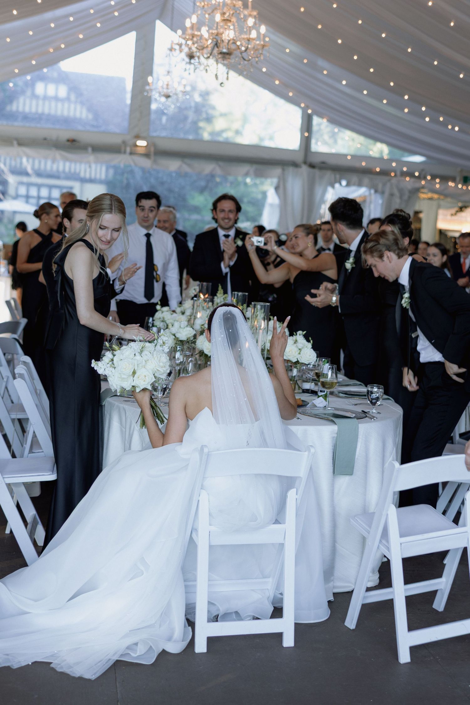 Elegant wedding reception with white decor, chandeliers and guests celebrating inside tent.