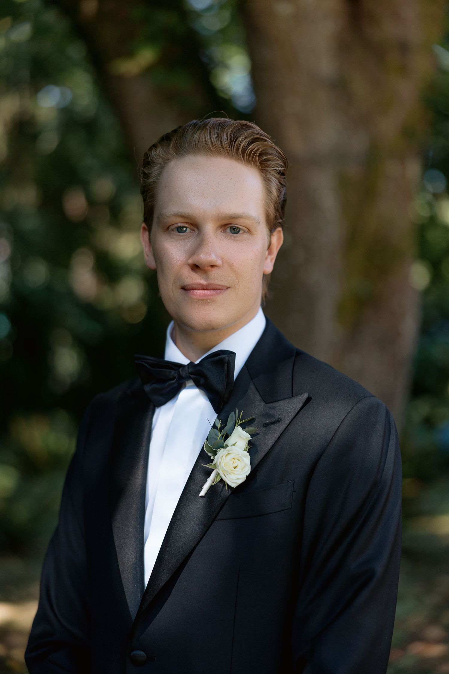 Groom in black tuxedo with white boutonniere poses for formal portrait outdoors.