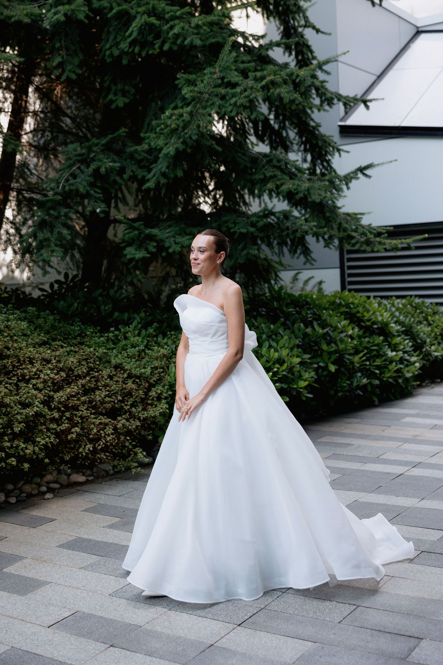A stunning white wedding gown with a flowing ballgown skirt photographed against greenery and modern architecture.
