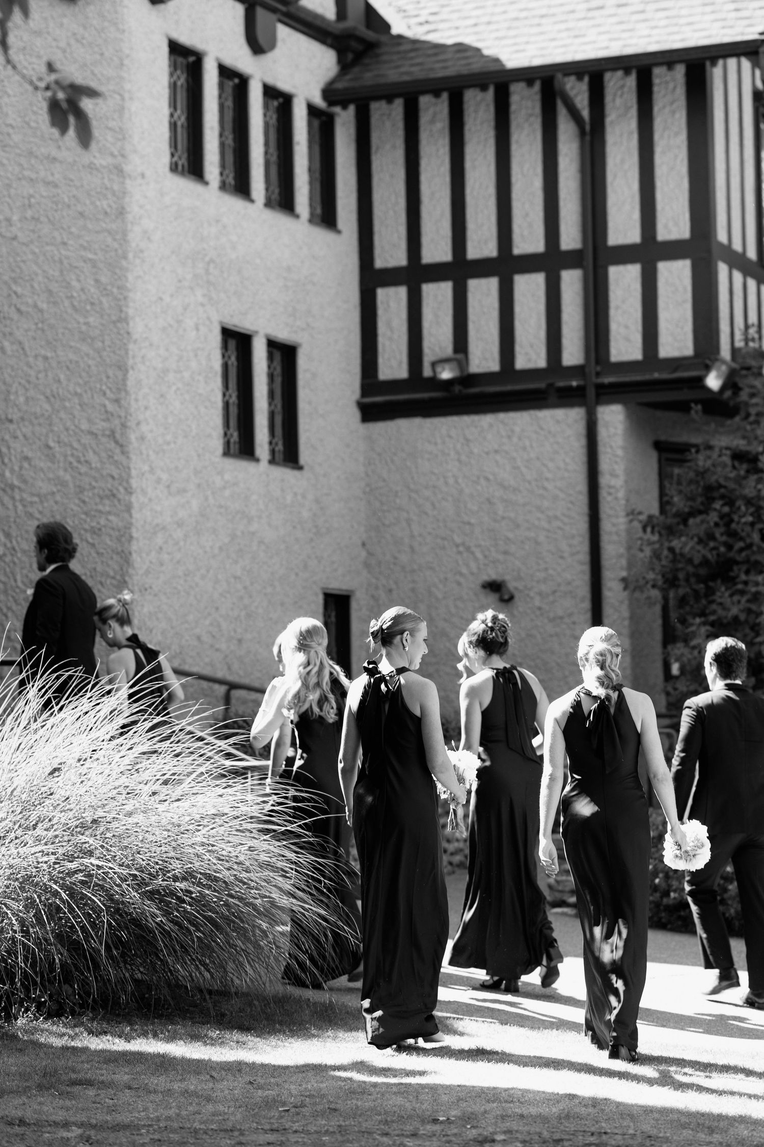 Wedding party members in formal black attire walk along Tudor building exterior.