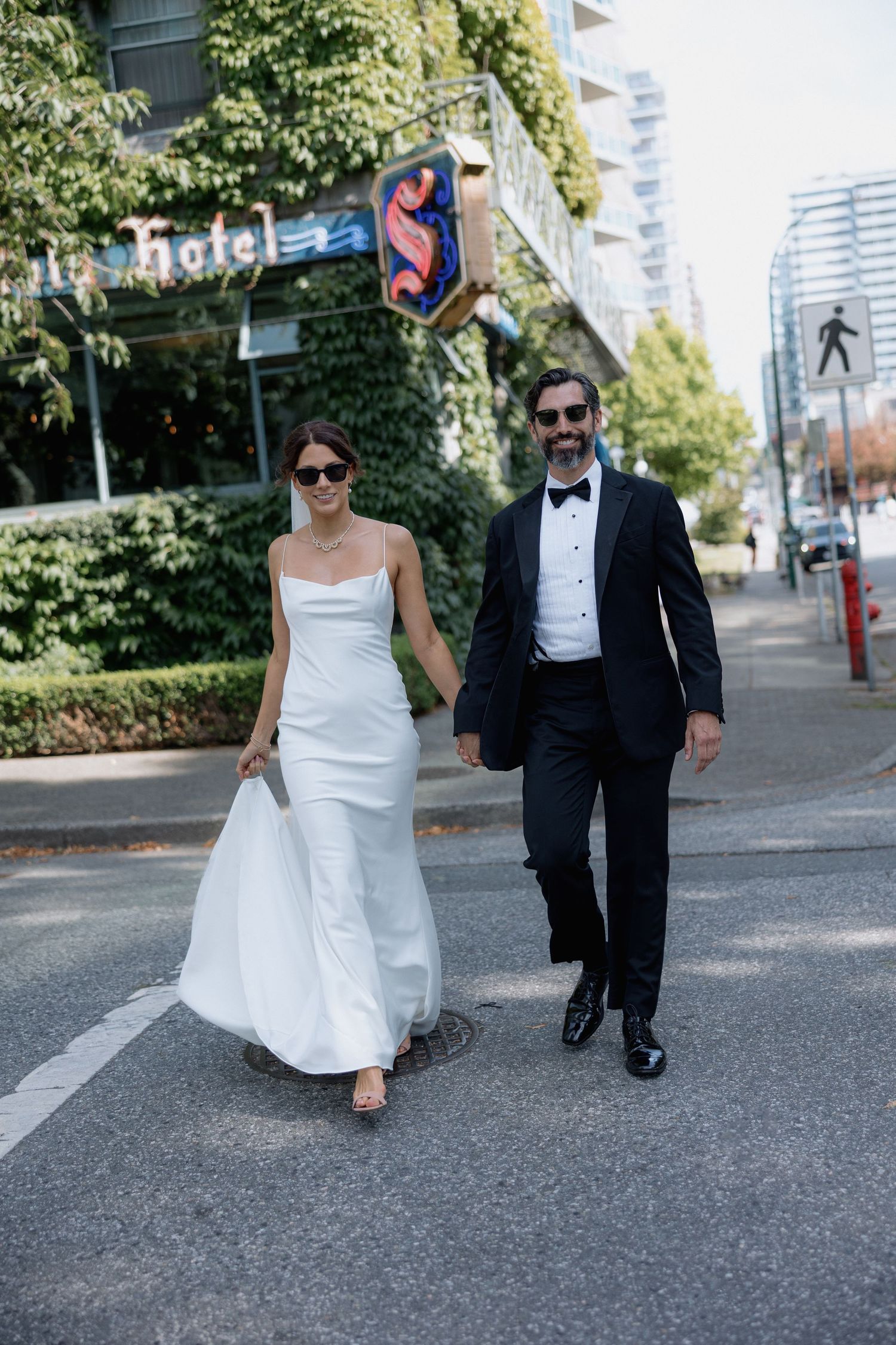 A couple walking on a city street, with the bride in a white gown and groom in a black tuxedo with bow tie in front of the Sylvia Hotel