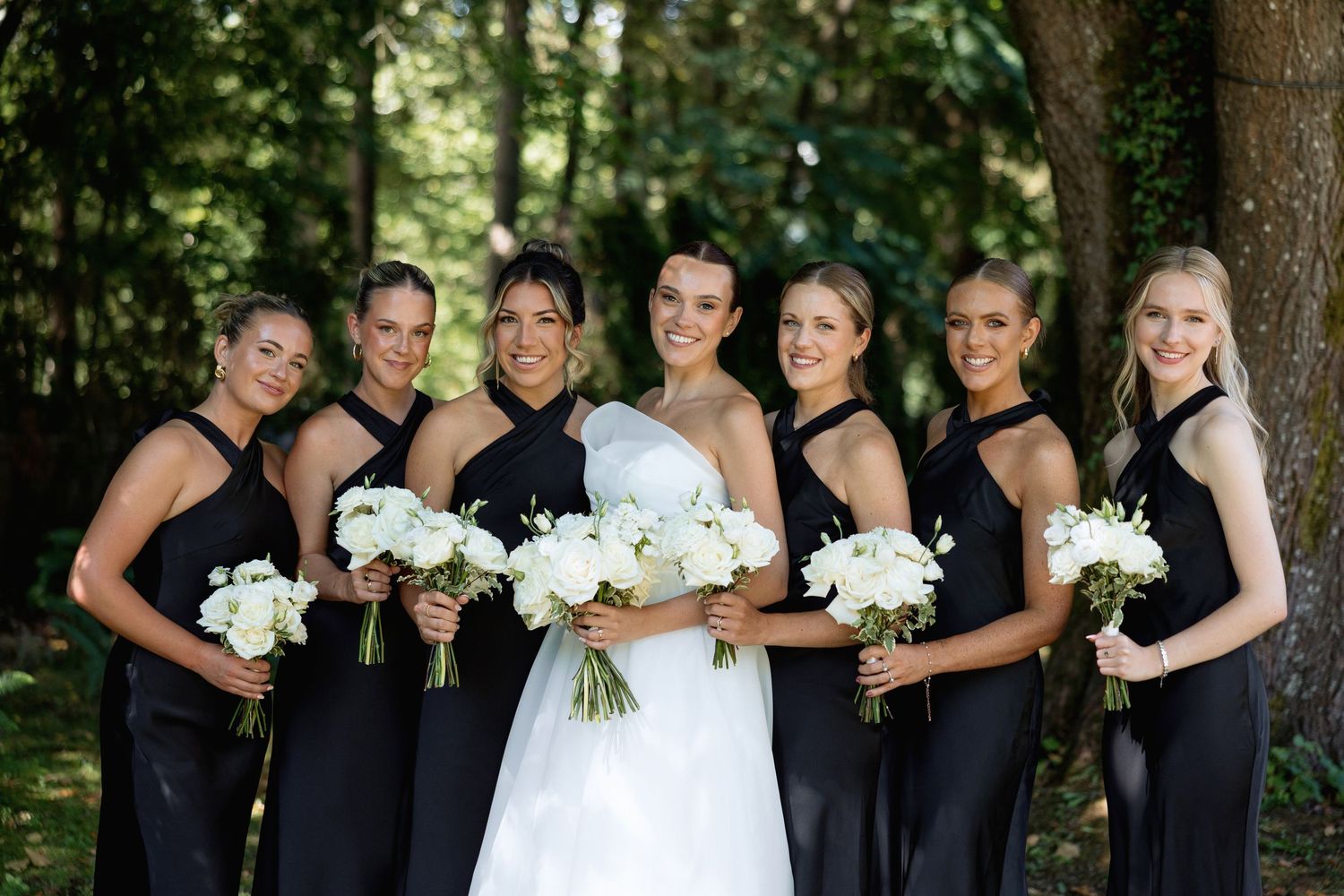 Bridesmaids in navy dresses pose with bride holding white bouquets outdoors.