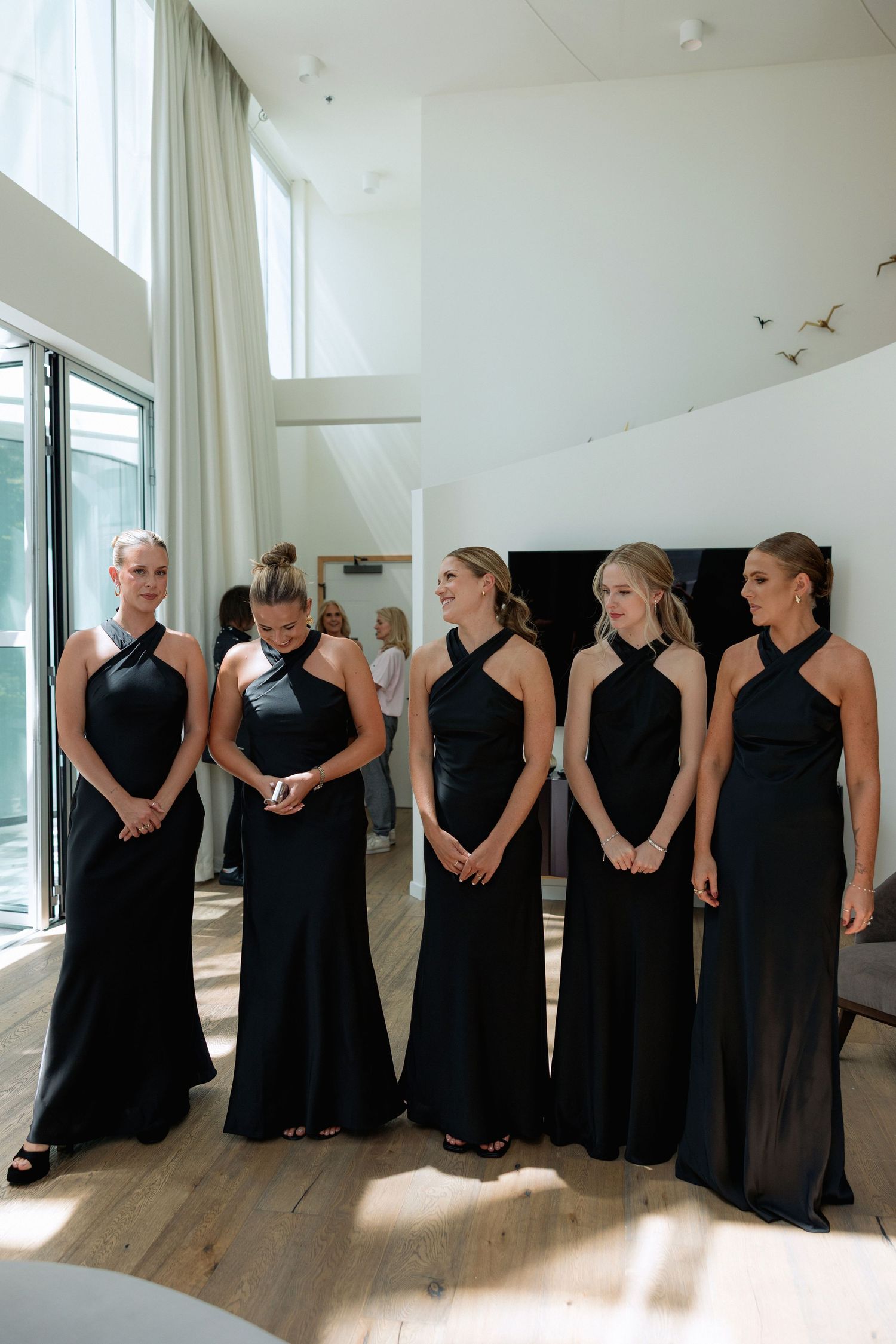 A line of people in black formal dresses stand against a white wall in a bright room.