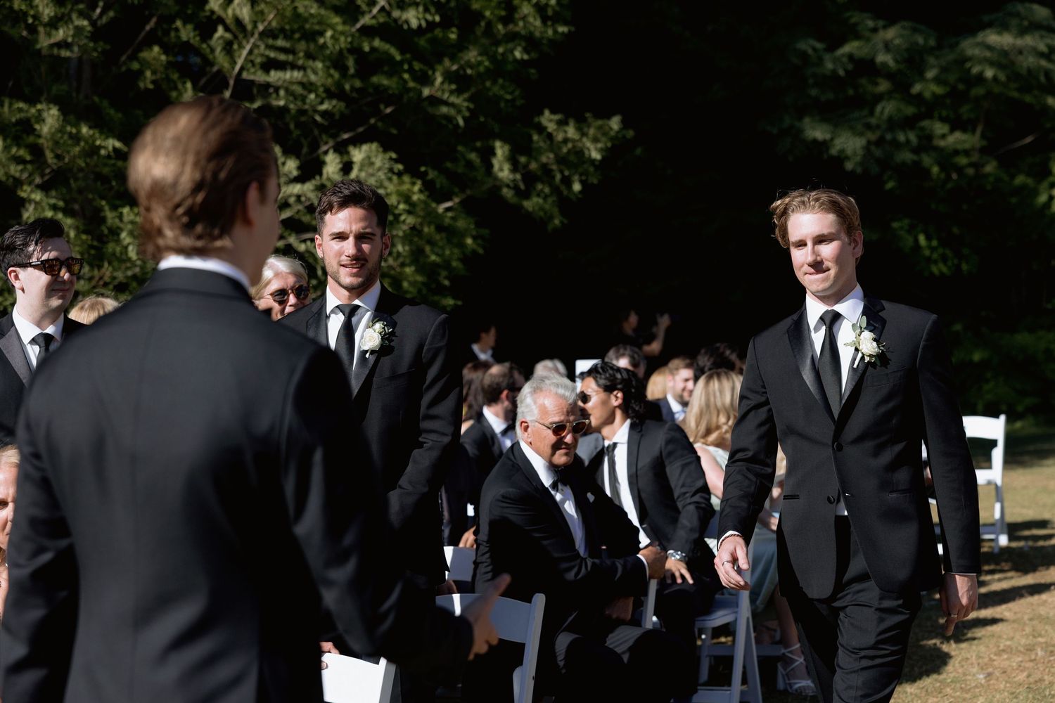 Formal outdoor wedding ceremony setup with guests in dark suits mingling before the event.