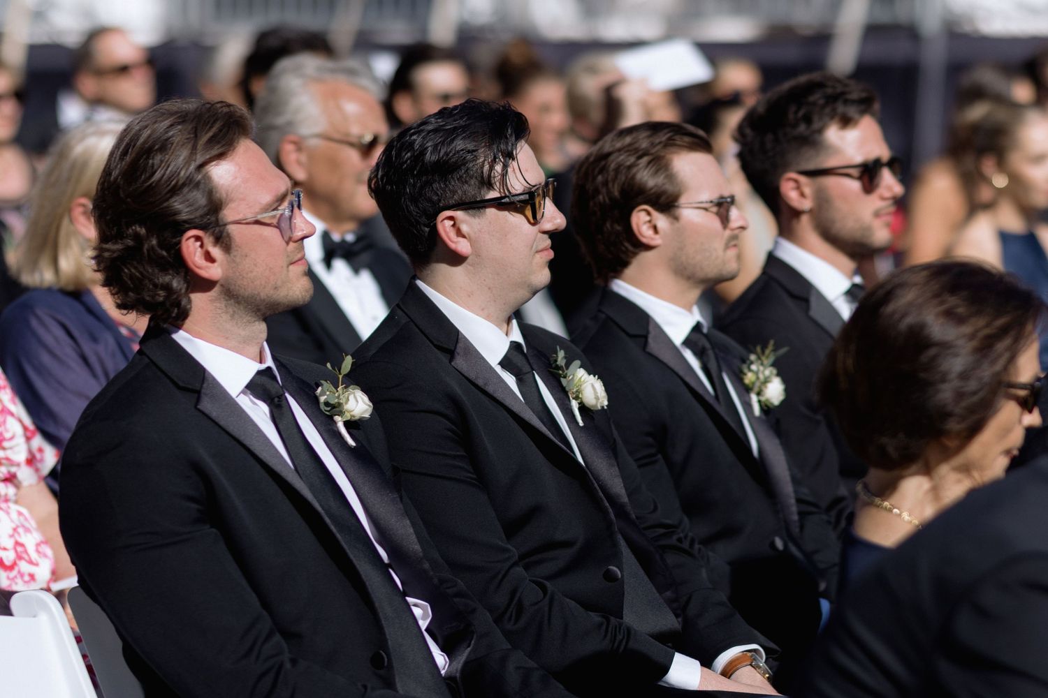 Row of wedding guests in formal black attire seated during outdoor ceremony.