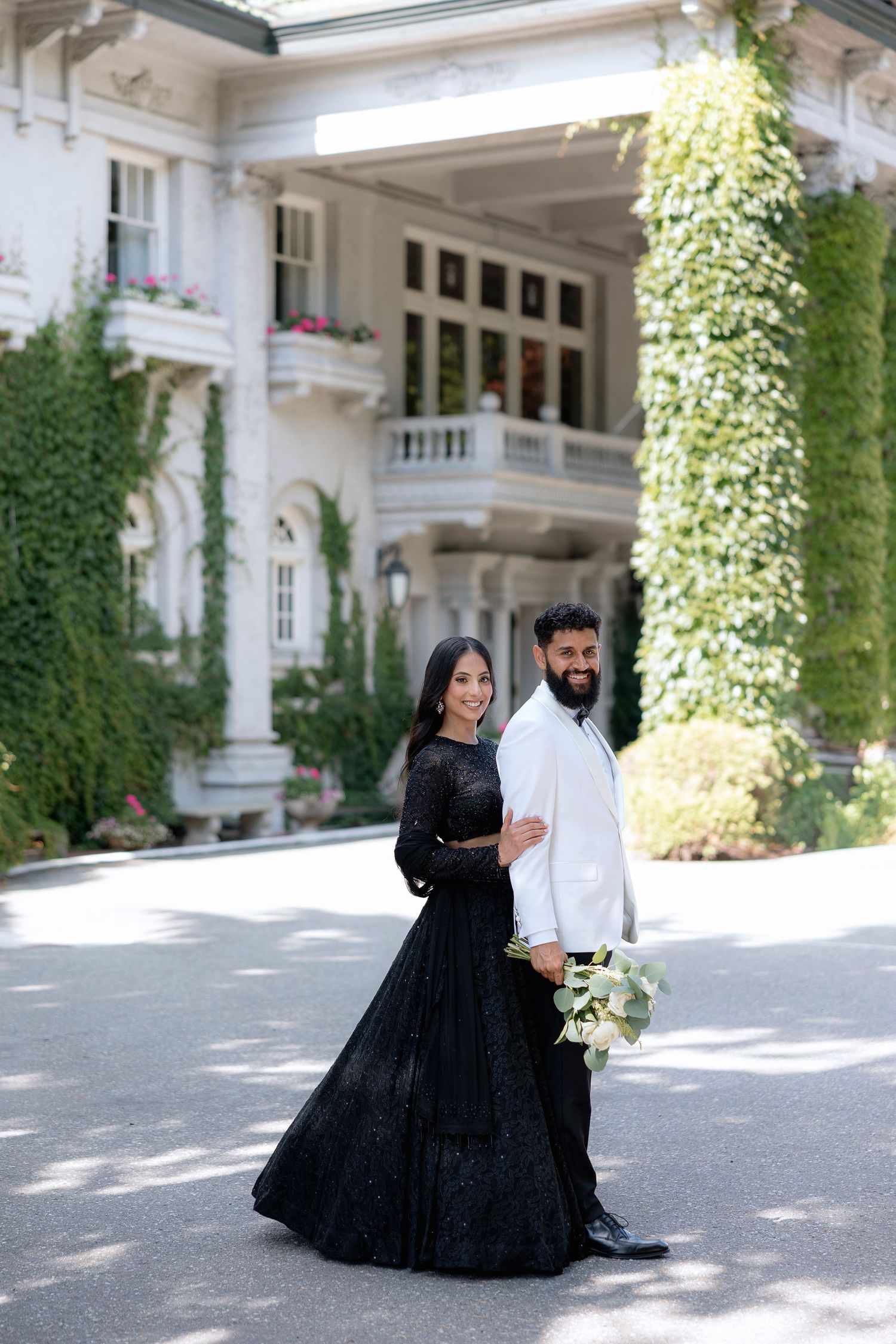 A couple in formal attire dances gracefully on a sunlit patio with white columns and climbing ivy in the background.