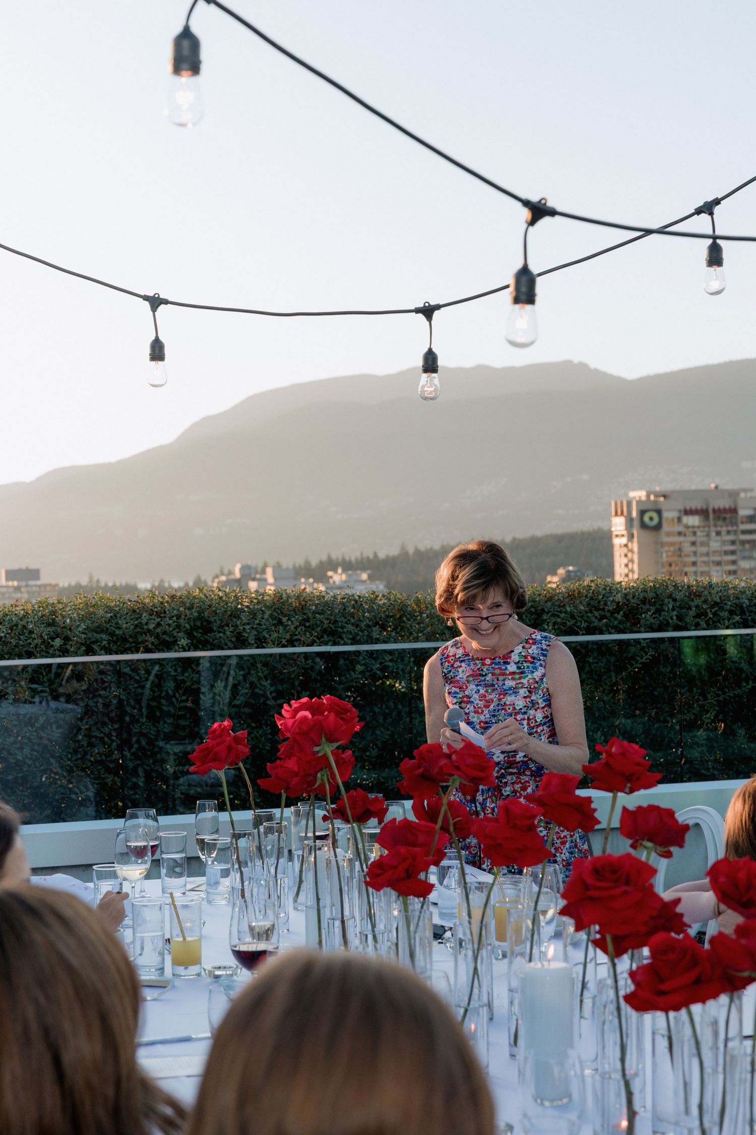 Outdoor dining setup with string lights and red flowers against a mountain sunset view.