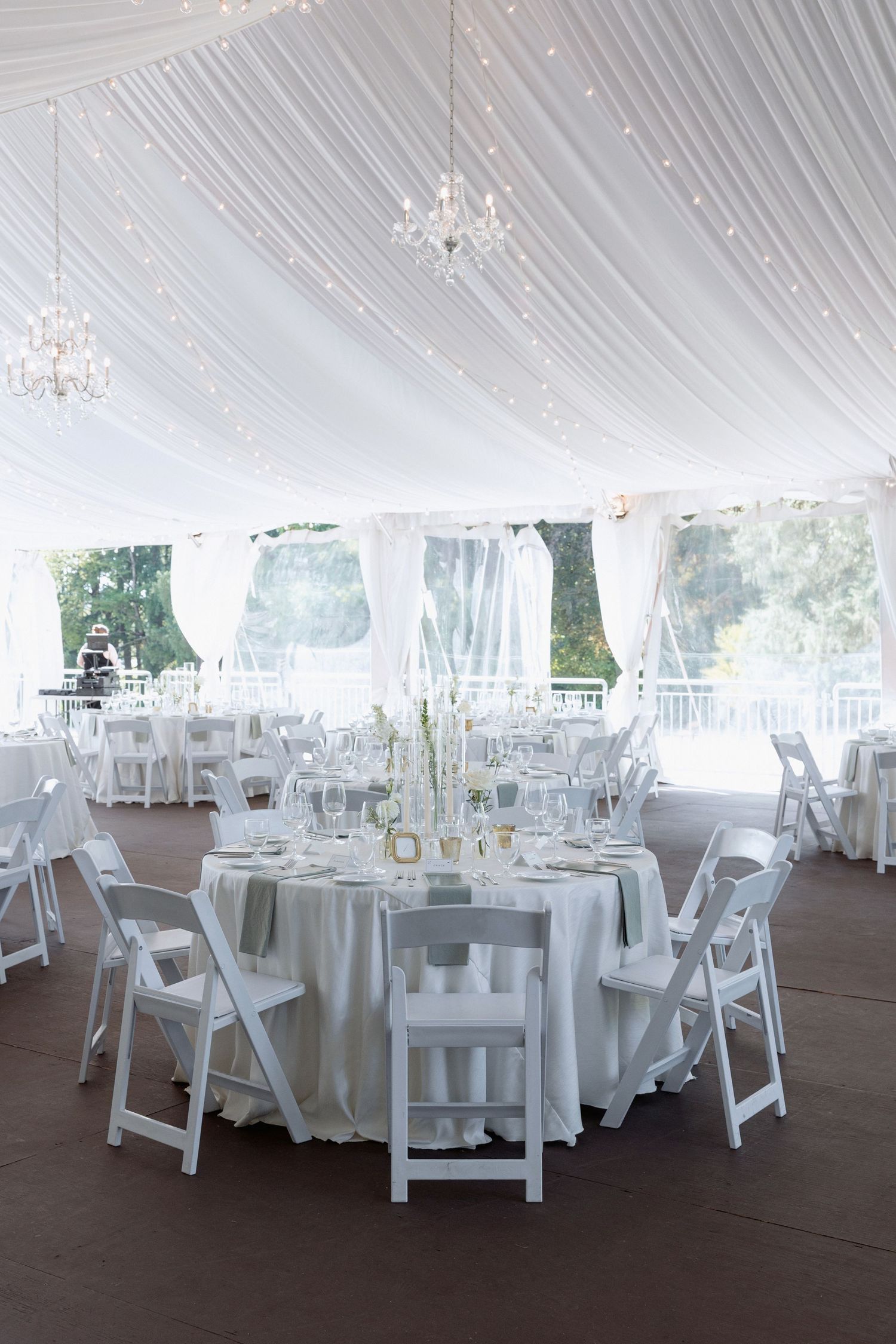 Wedding reception setup with round tables and white chairs under a draped tent with chandeliers.