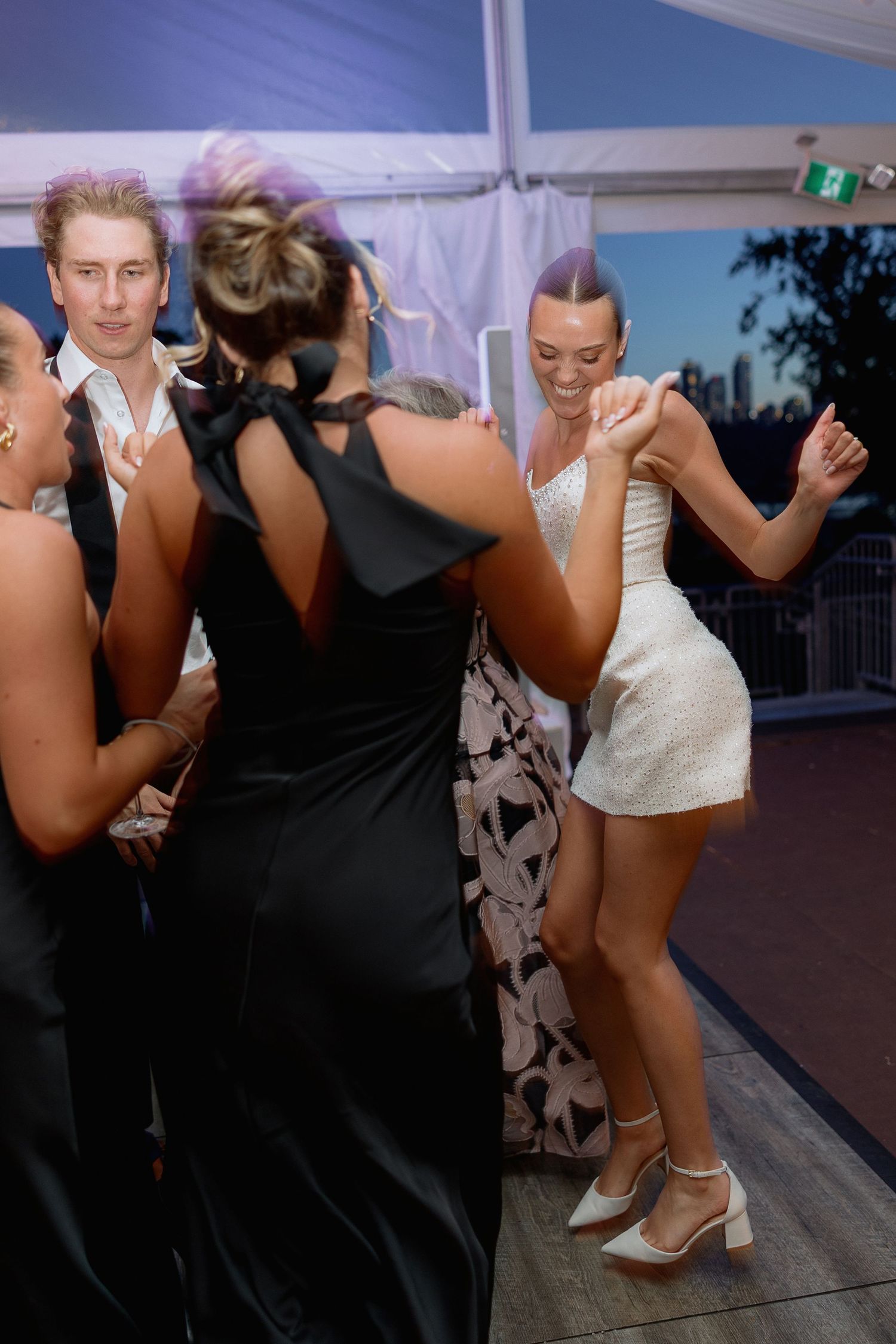 Wedding guests dance energetically on a wooden dance floor under string lights at a reception.
