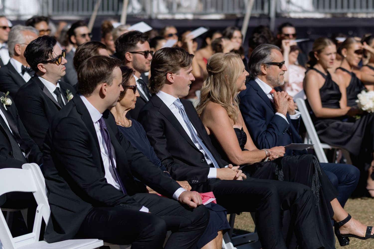 Formally dressed wedding attendees sitting in rows during an outdoor ceremony.